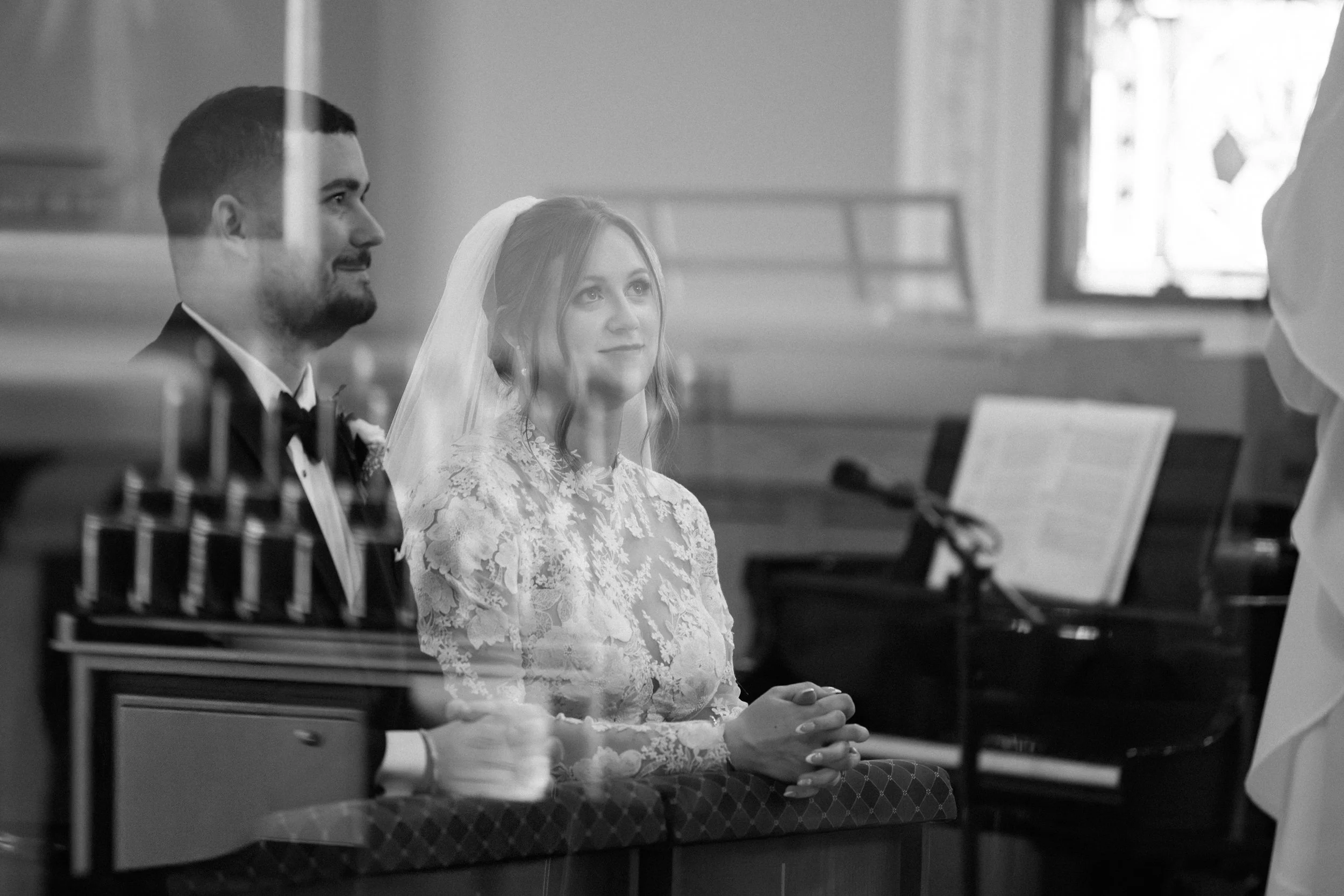 A couple at a wedding ceremony, with the groom wearing a tuxedo and the bride in a lace dress and veil, sitting at the altar inside a church.