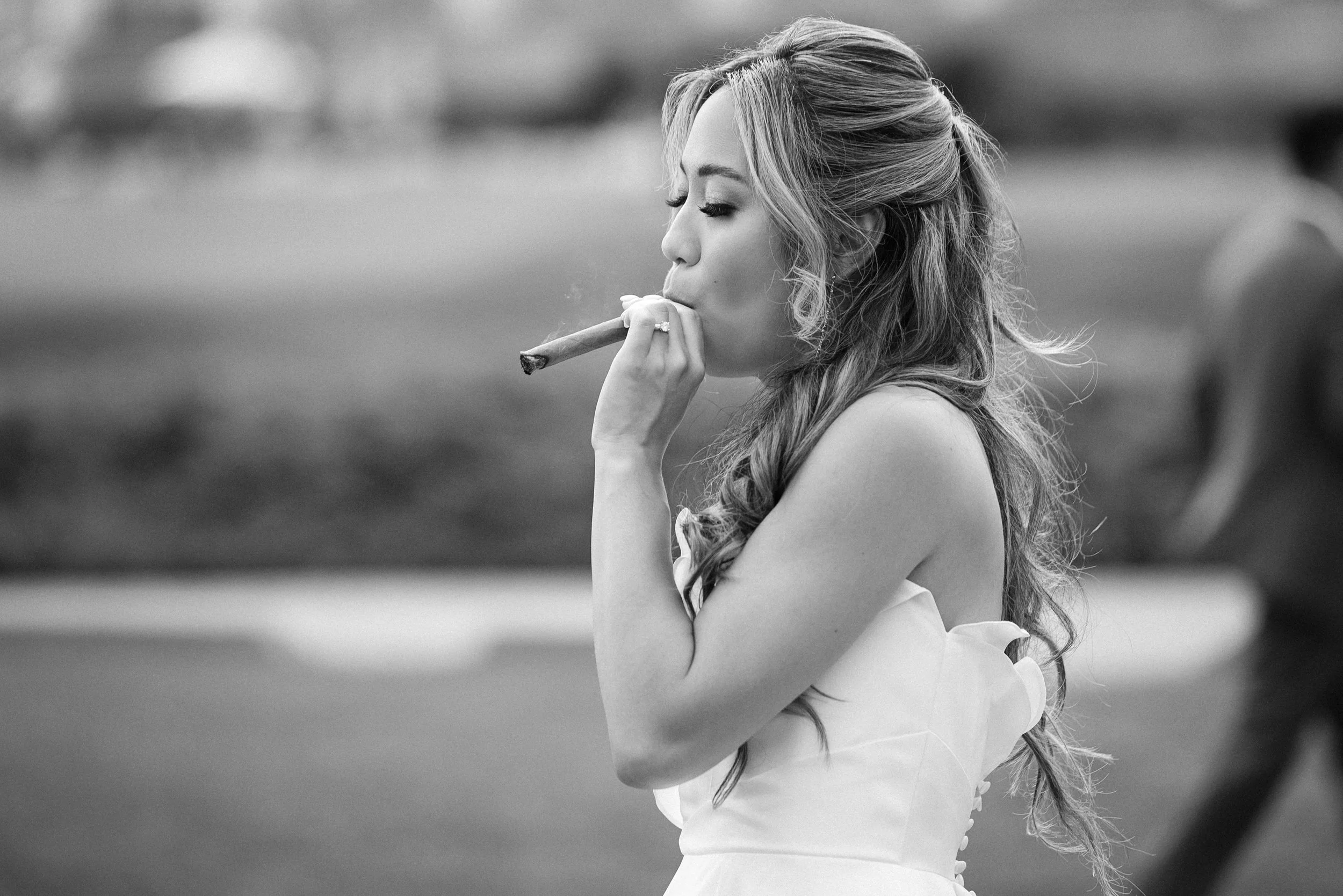 A woman with long, wavy hair in an updo, wearing a strapless white dress, is smoking a cigar outdoors.