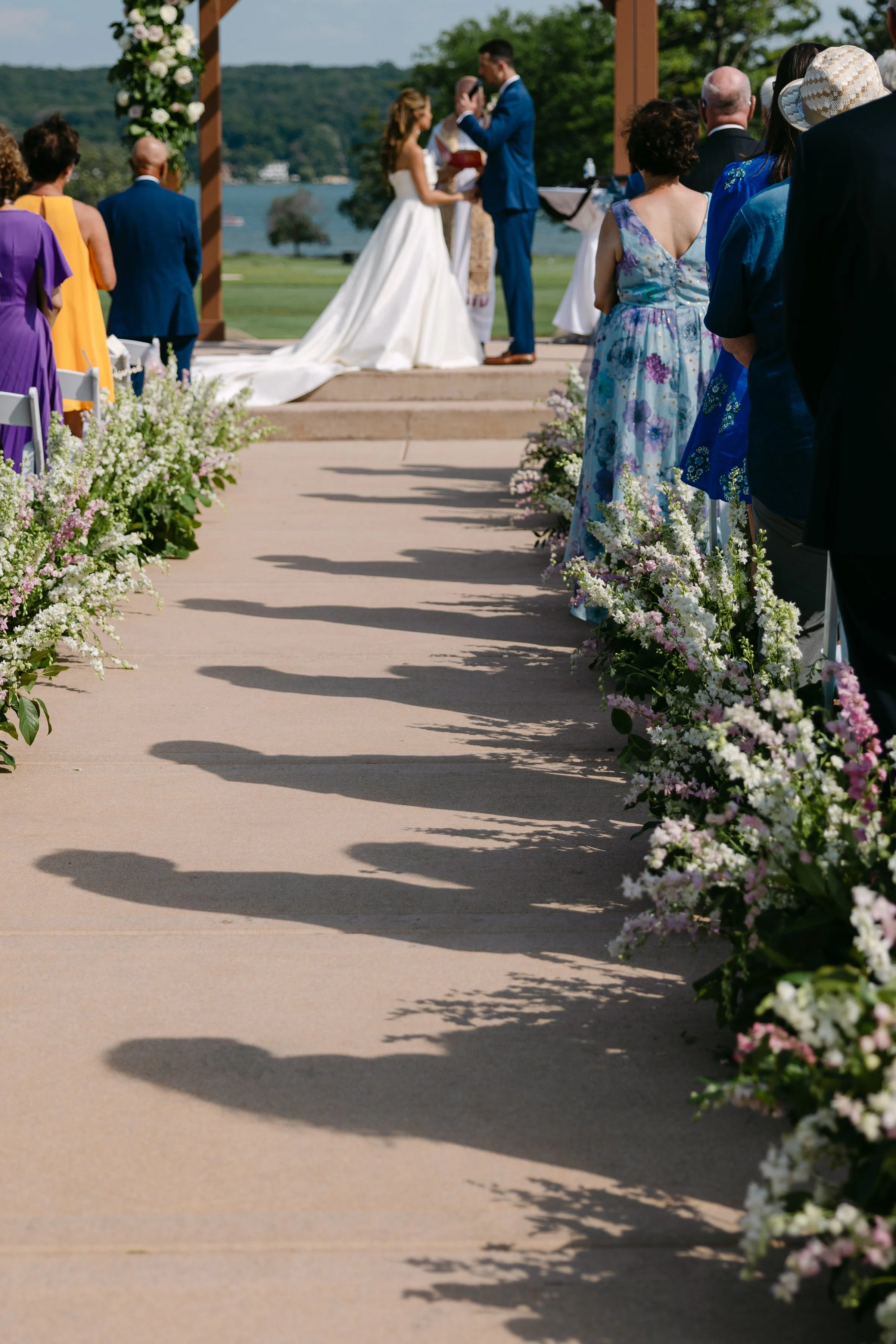 Outdoor wedding ceremony with a couple exchanging vows under a decorated arch, guests seated beside flower arrangements along the aisle, water and trees in the background.