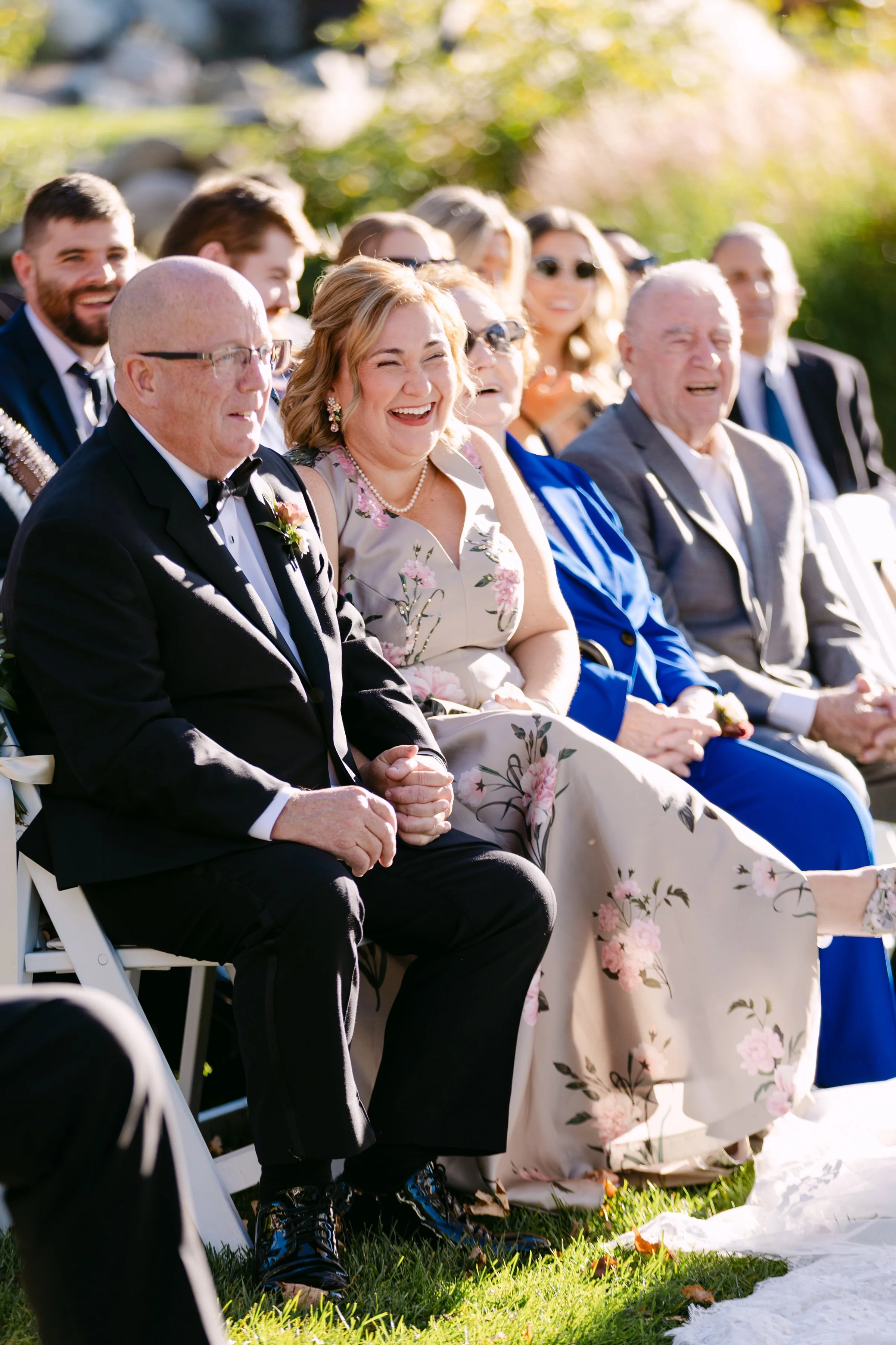 Group of people sitting outdoors at a wedding, smiling and laughing in sunlight.
