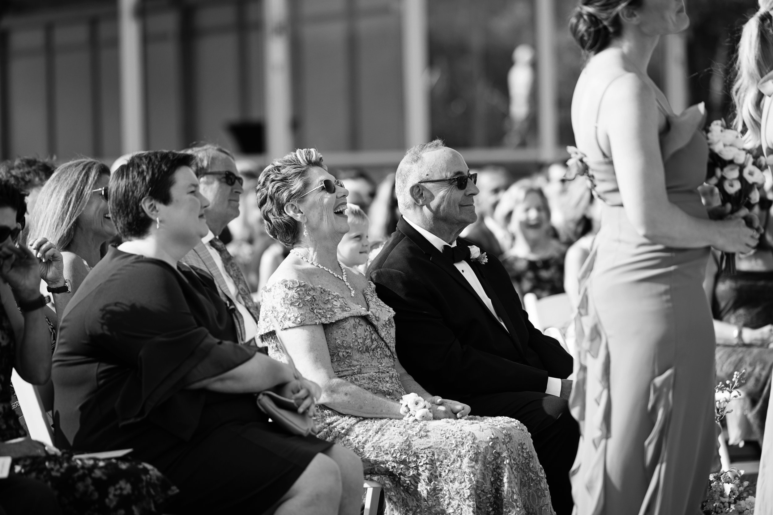 Black and white photo of people sitting and watching a wedding ceremony, including an elderly woman laughing and an elderly man with sunglasses.