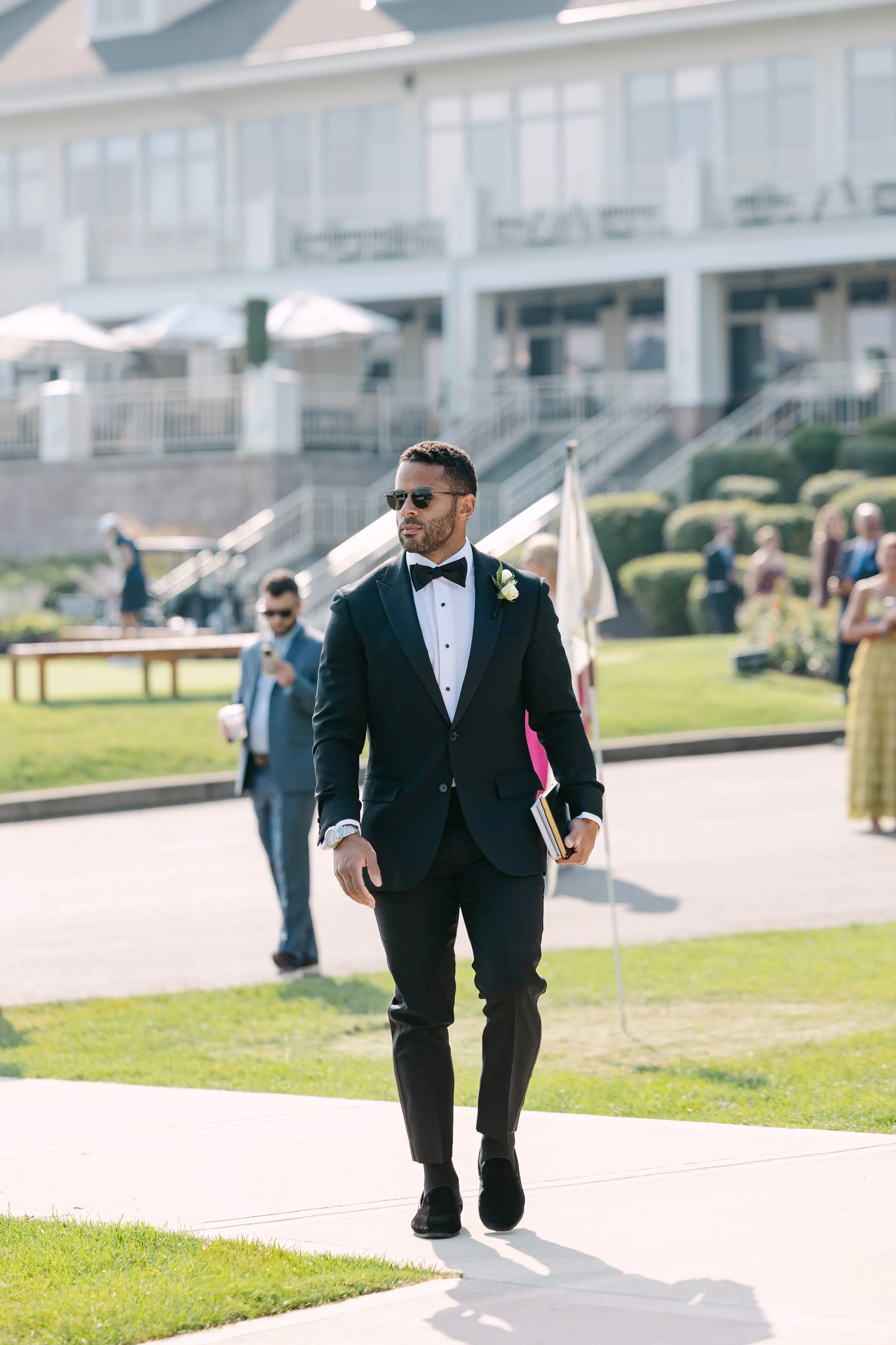A man in a black tuxedo, sunglasses, and a bow tie walking outdoors at a formal event, holding a book. There are other people in the background, some dressed formally, with a building and grass area behind them.