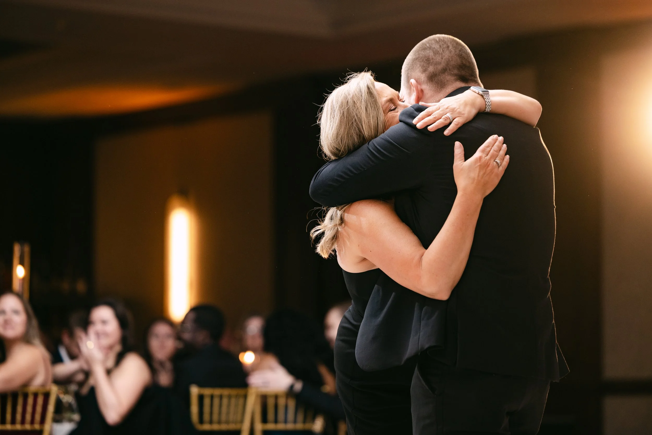 A woman and a man sharing a warm embrace at a formal event, with seated guests in the background.