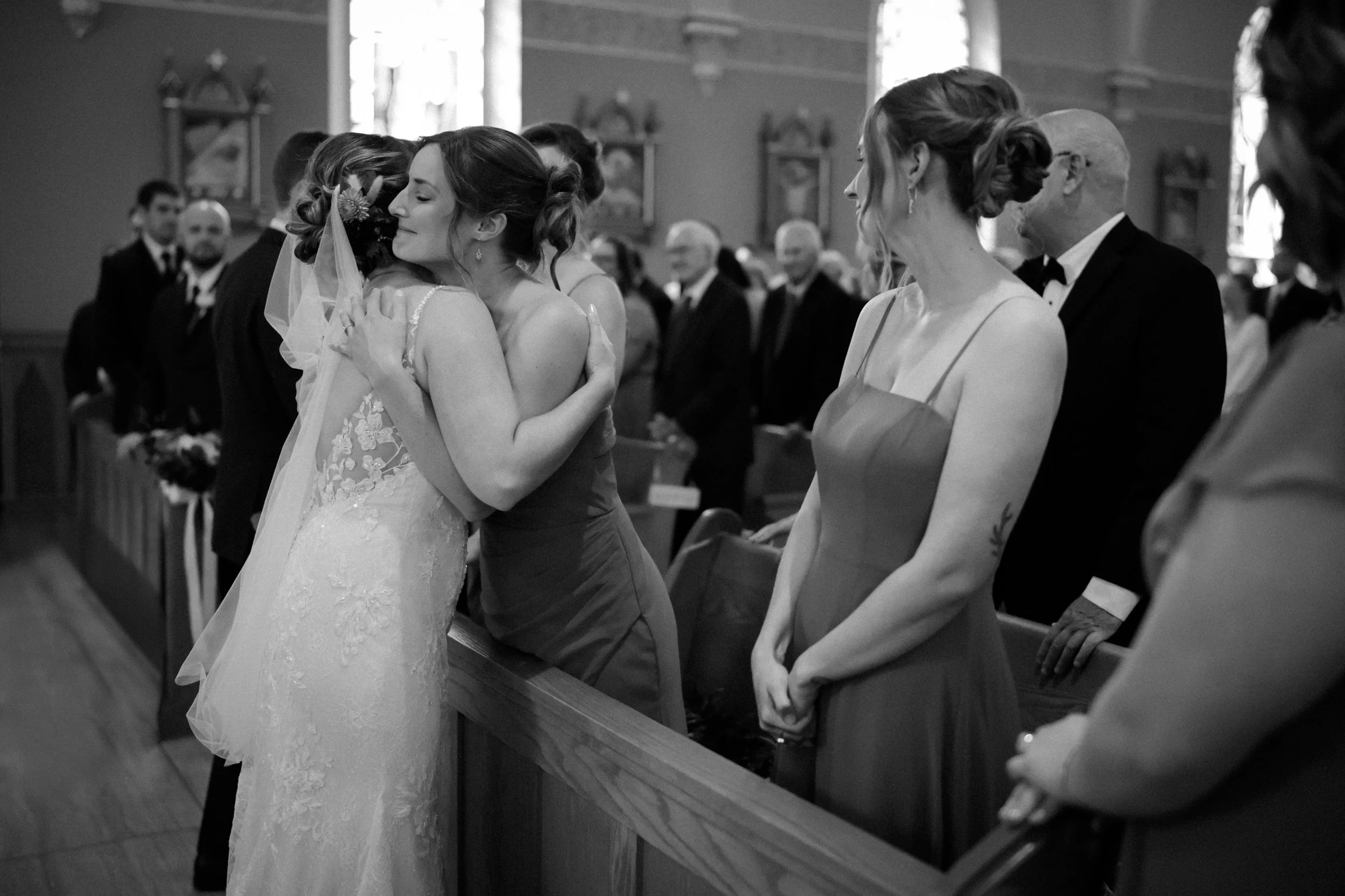 Two women hugging during a wedding ceremony inside a church, surrounded by friends and family, all dressed in formal attire.