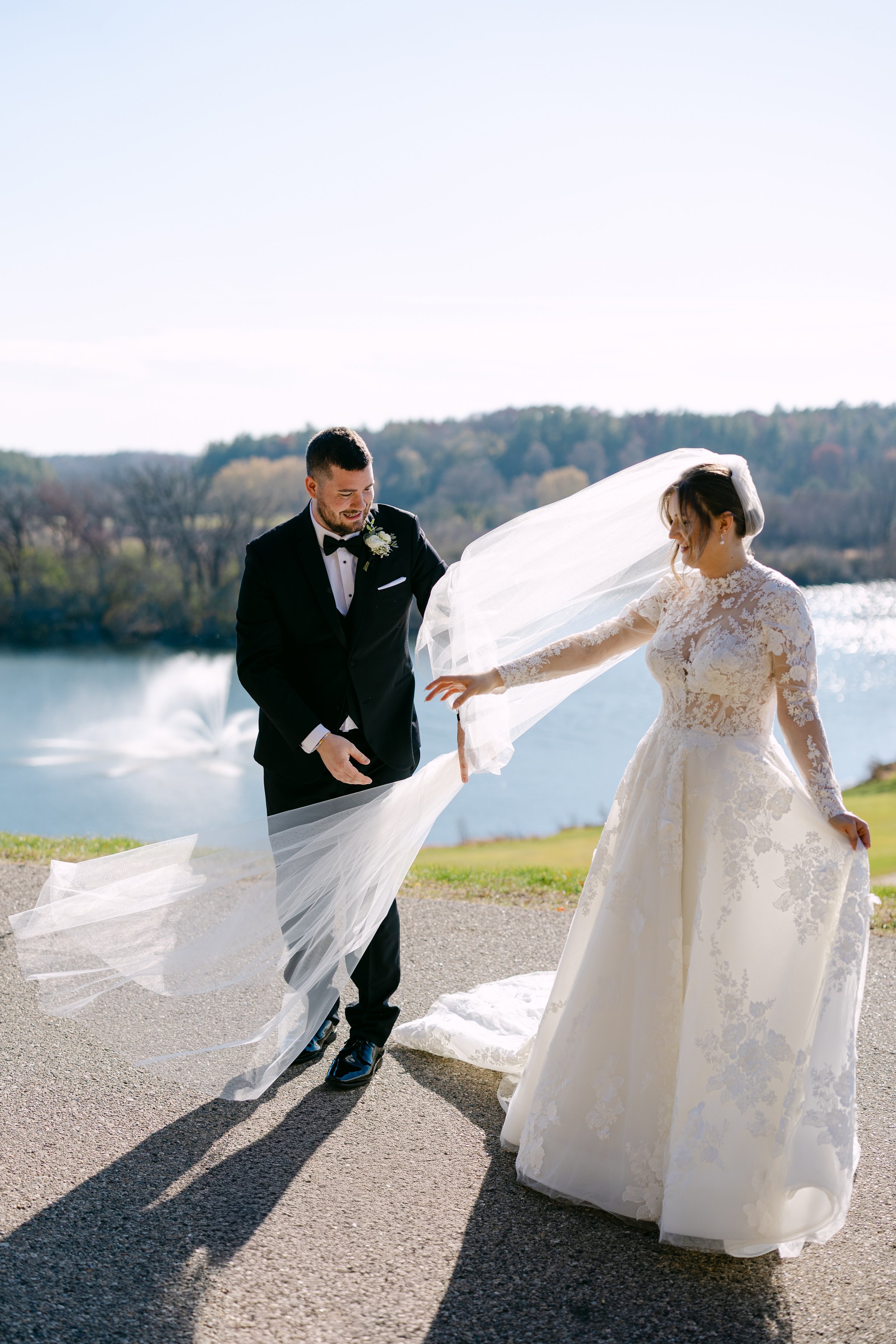 A bride and groom stand outdoors by a body of water, the bride holding her veil while the groom helps to adjust it.