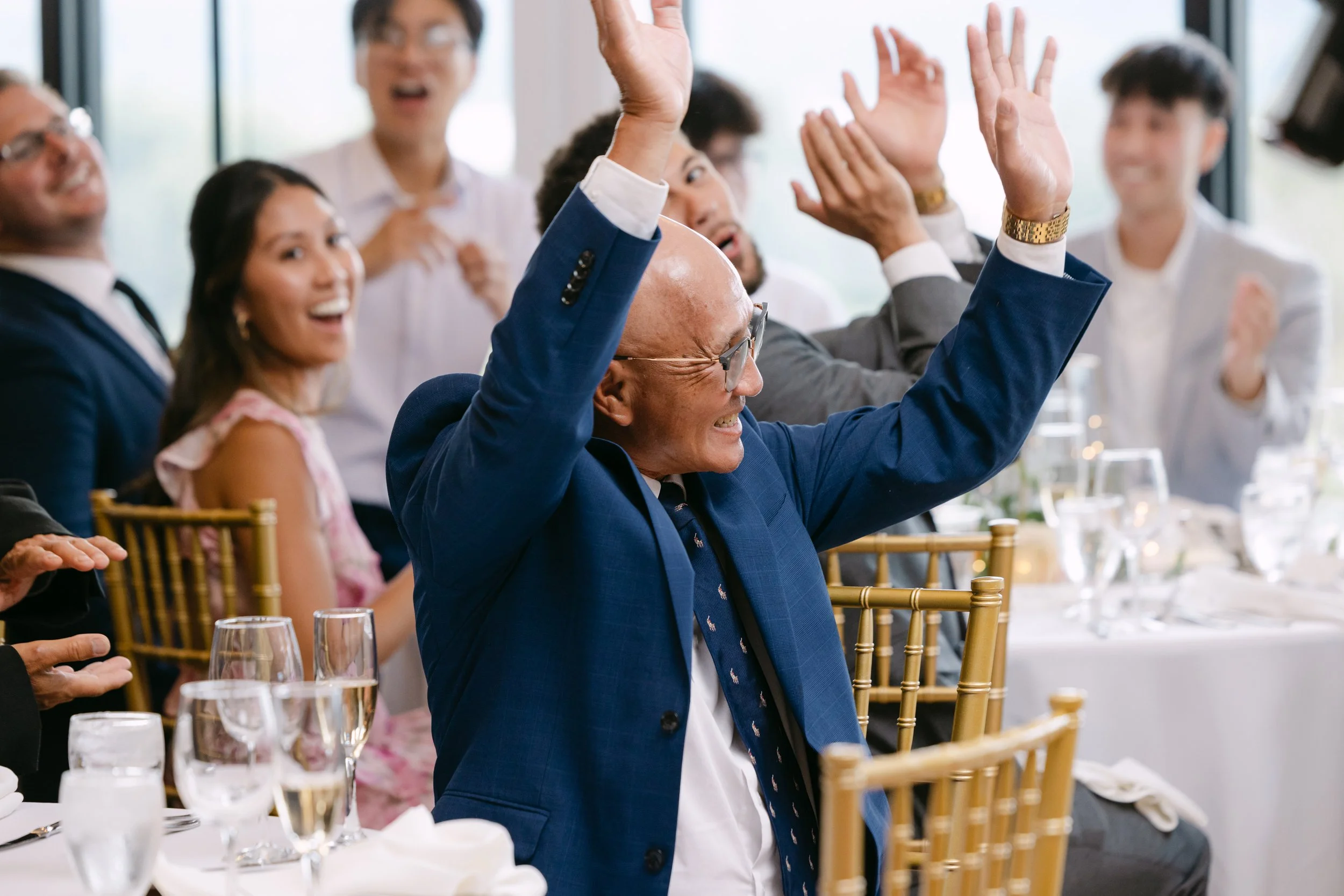A group of people at a celebration, with one man in a blue suit and glasses raising his hands and smiling, surrounded by others who are clapping and smiling at a formal event or party.
