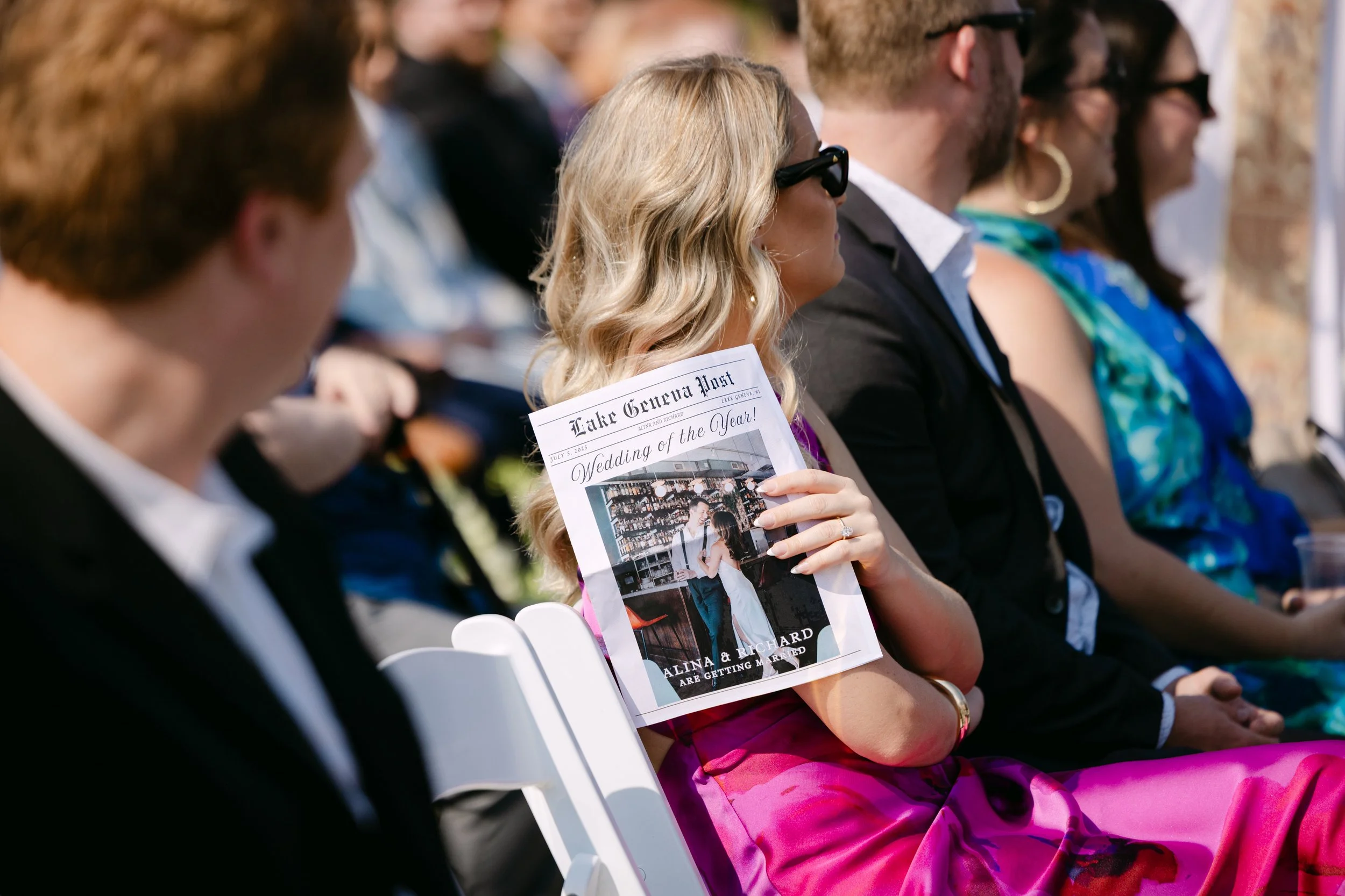 A woman in a pink dress wearing sunglasses is sitting in an outdoor wedding ceremony, holding a newspaper titled 'Lake Geneva Post' with a photo of a couple on the cover.