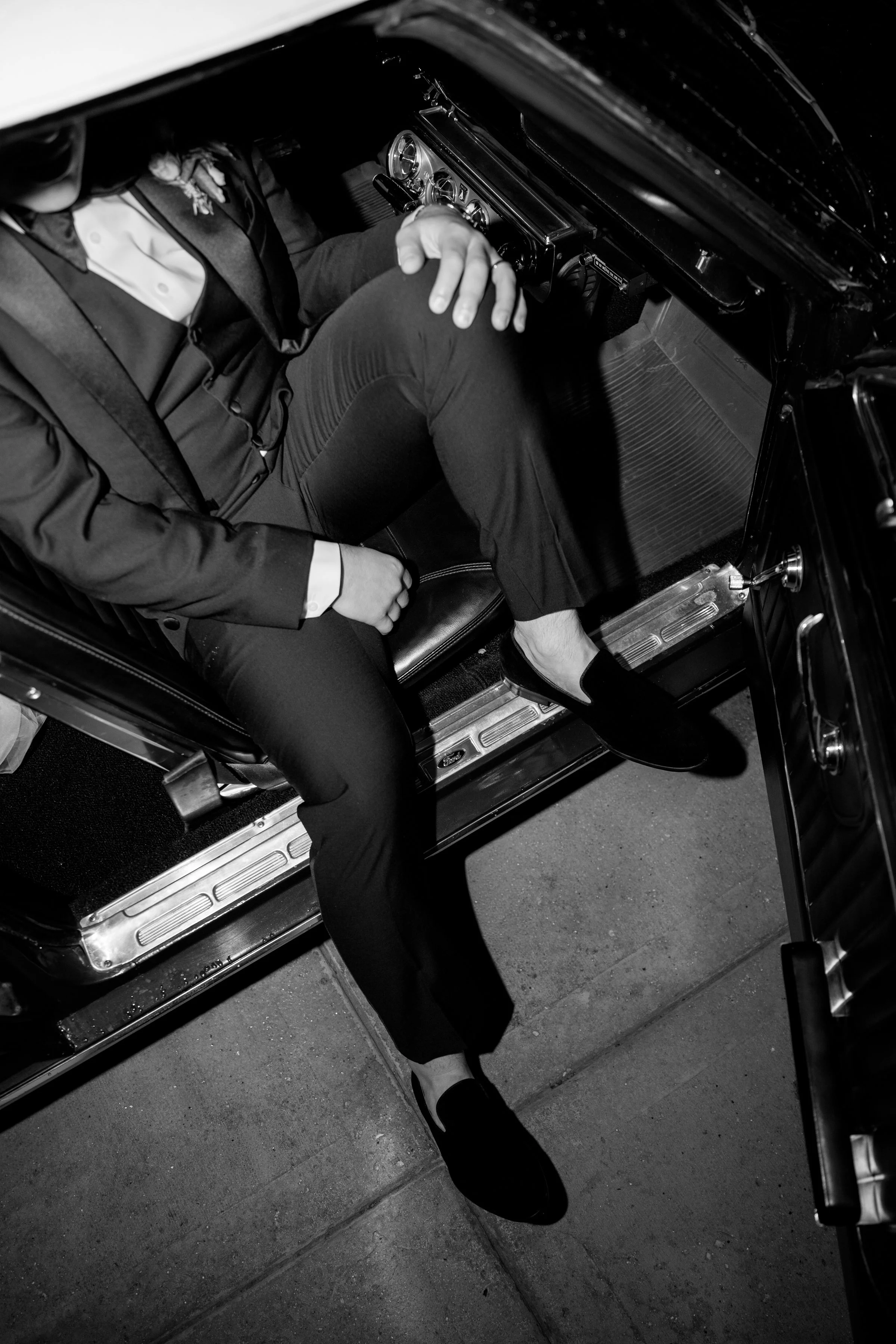 A person sitting inside a vintage car, dressed in formal attire, with one leg crossed over the other, wearing black shoes, and the car's interior in black and chrome.