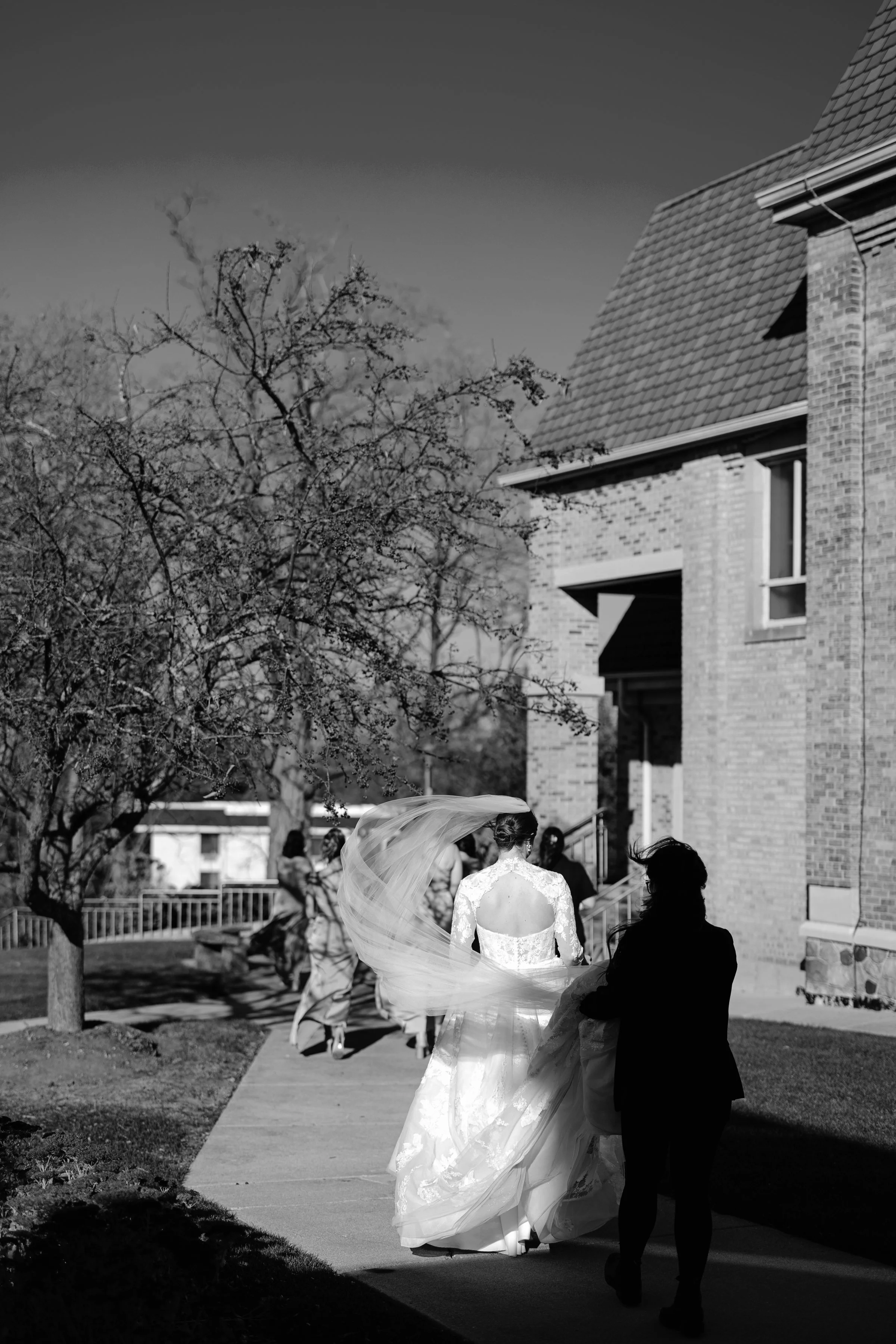 A bride walking outdoors on a sidewalk, surrounded by guests, with a woman carrying her dress, in front of a brick building and leafless trees.