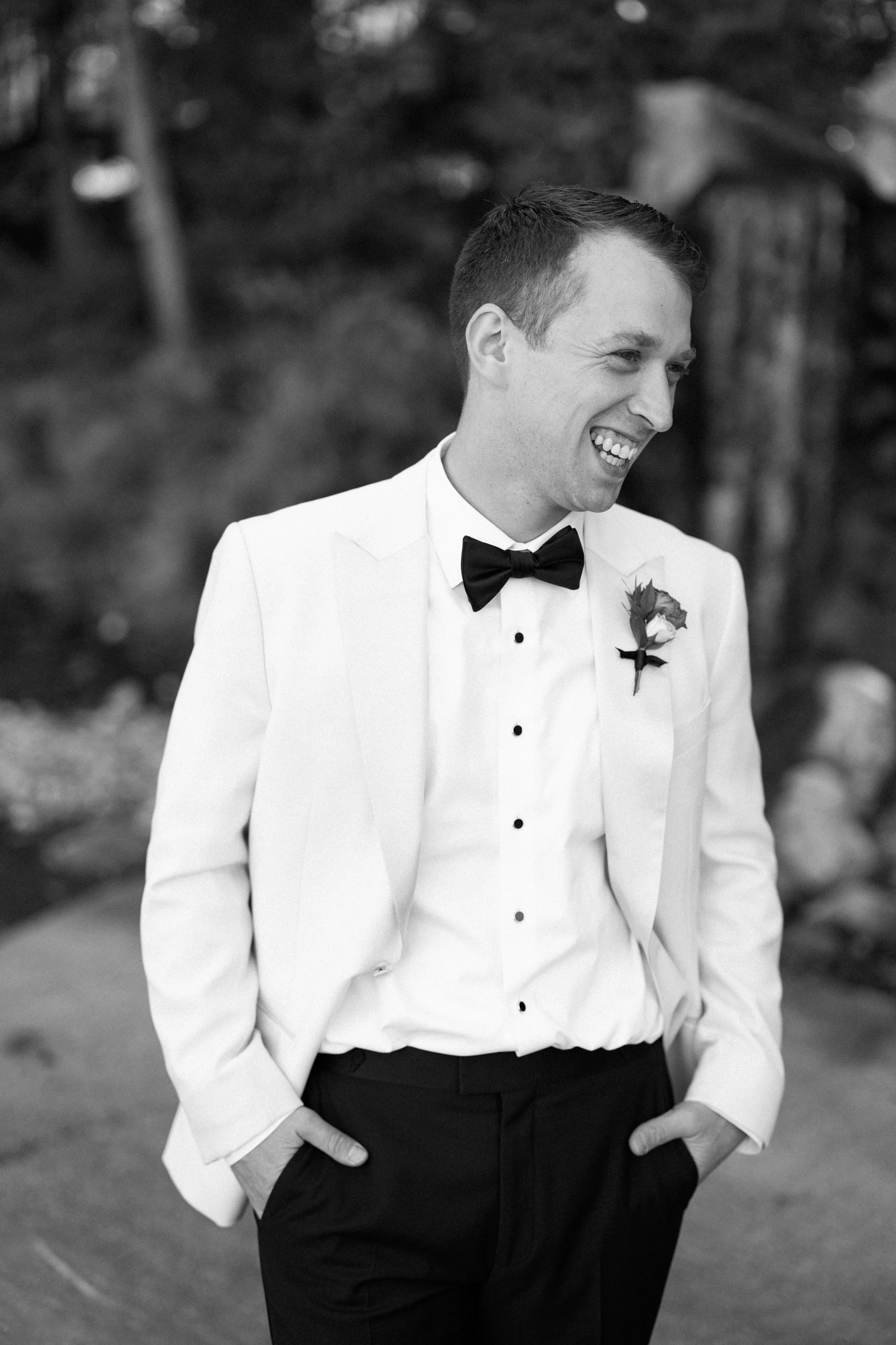 A man wearing a white tuxedo with a black bow tie and boutonniere, smiling with hands in pockets outdoors.