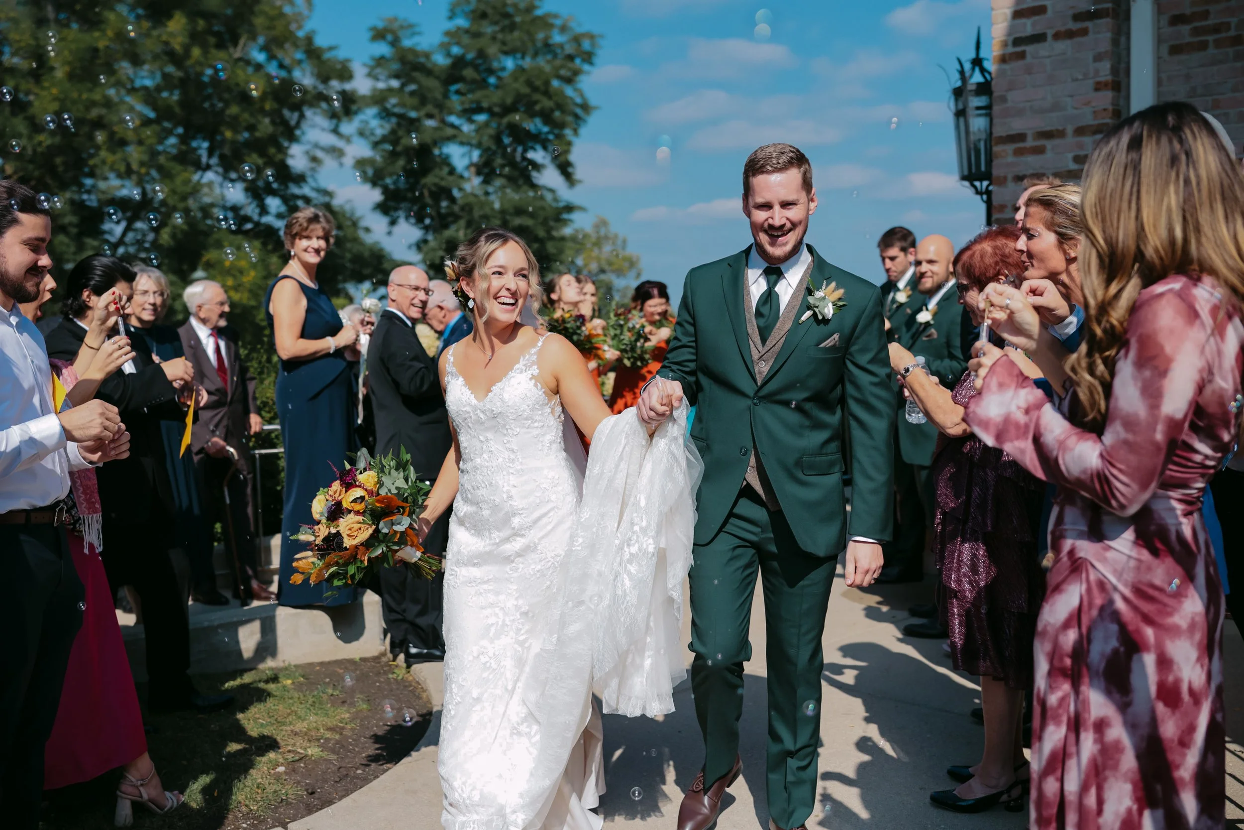 A bride and groom walking hand in hand through a crowd of celebrating guests outdoors on a sunny day during a wedding ceremony.