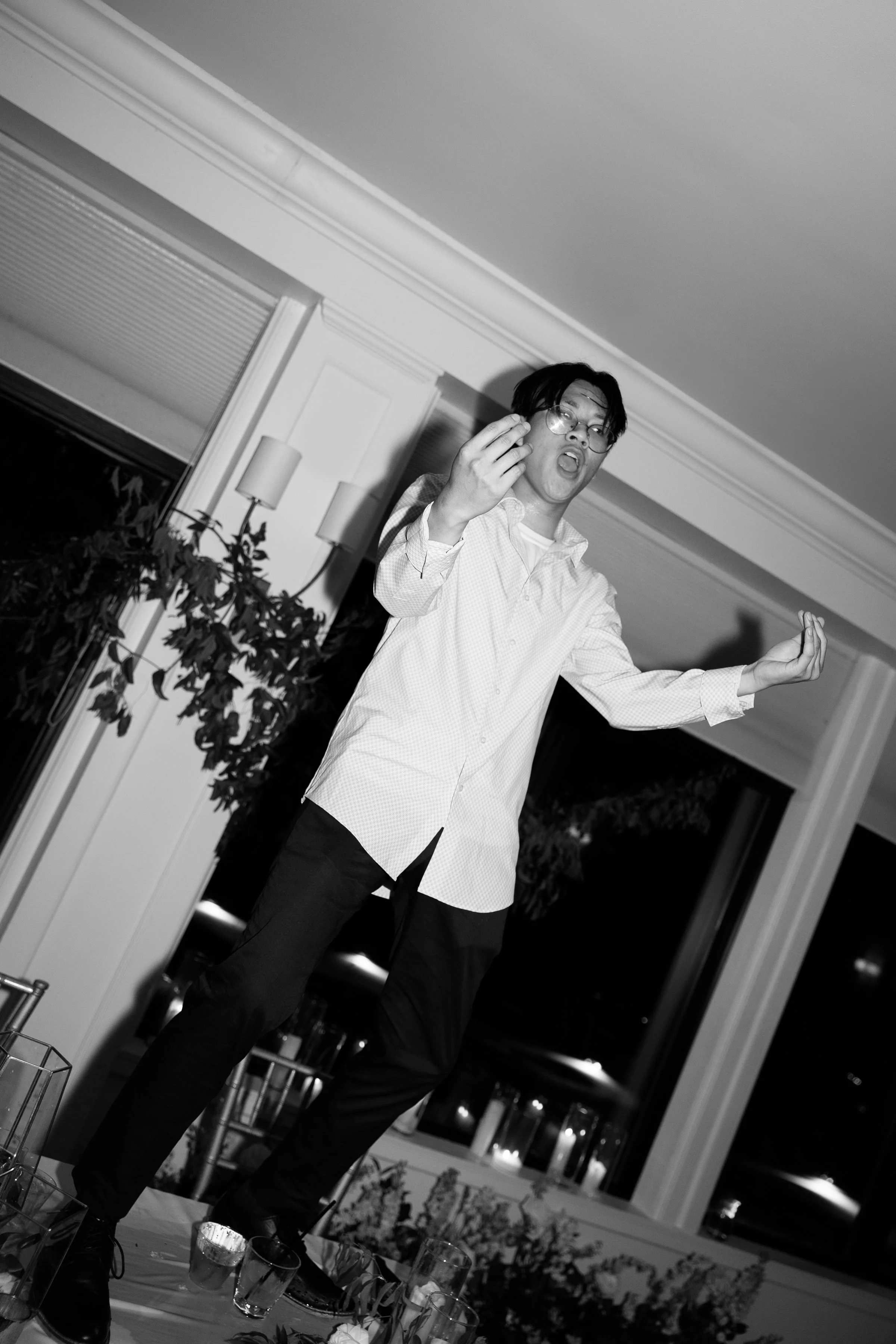 A man with glasses and dark hair in a white shirt is standing on a table, gesturing with his hands, in a room with large windows and evening outdoor scenery.