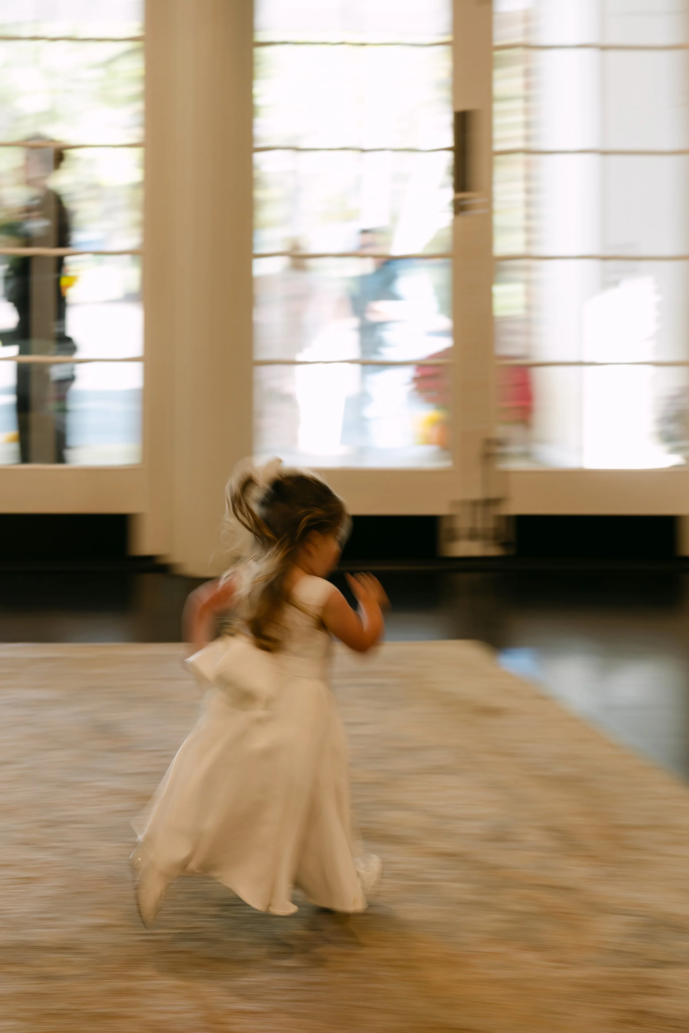 A young girl in a white dress is spinning or running inside a building with glass doors, with people and outdoor scenes visible behind the doors.