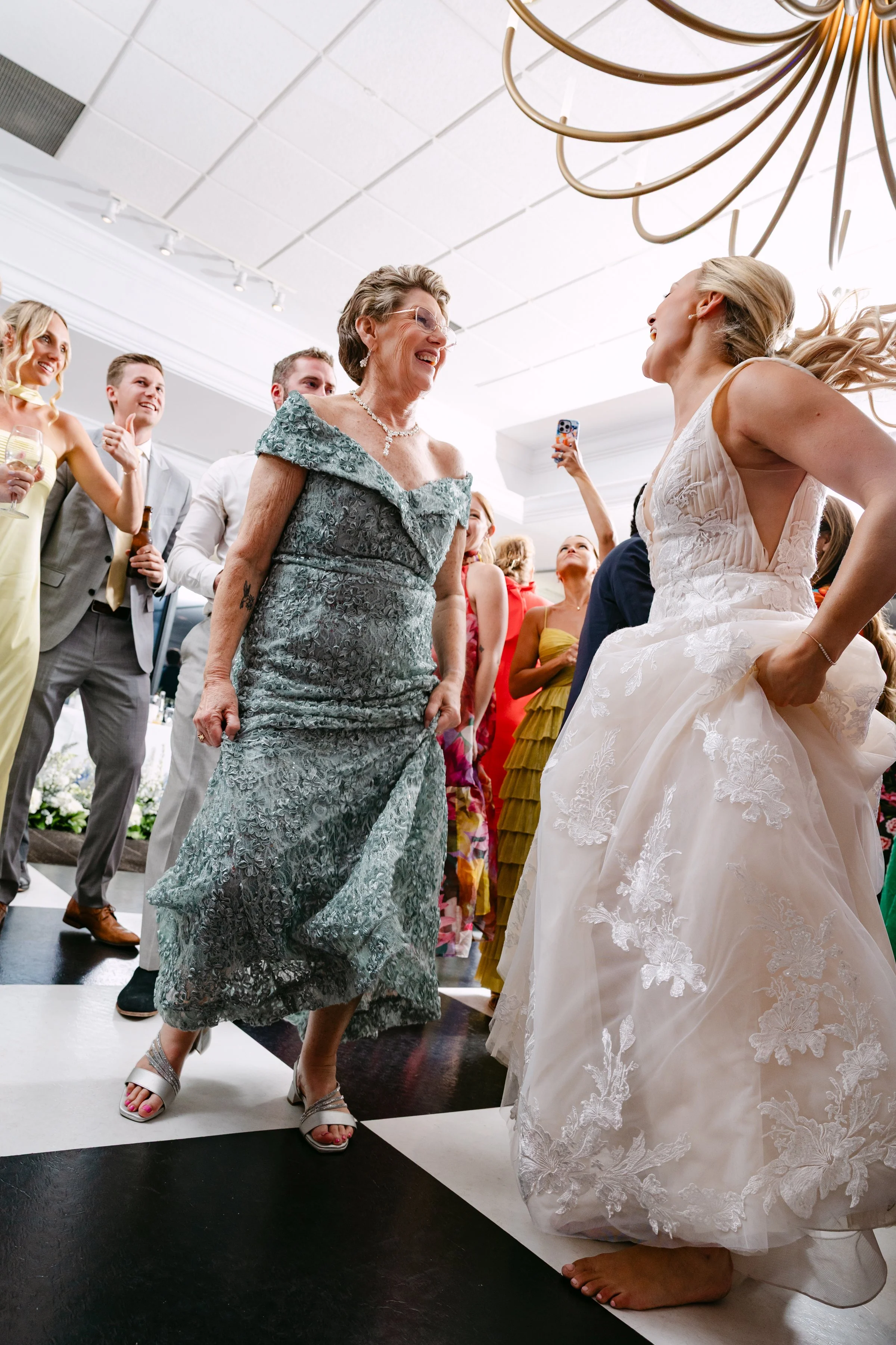 Women dancing at a wedding reception, with a bride in a white gown and older woman in a silver dress smiling at each other.