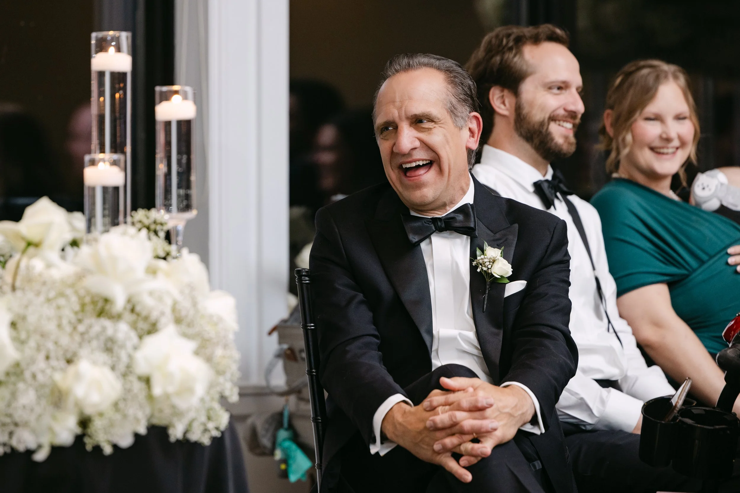 Group of people sitting at a formal event, with the focus on a man in a tuxedo smiling and sitting with his hands clasped, with two other people smiling in the background, near floral decorations and candles.