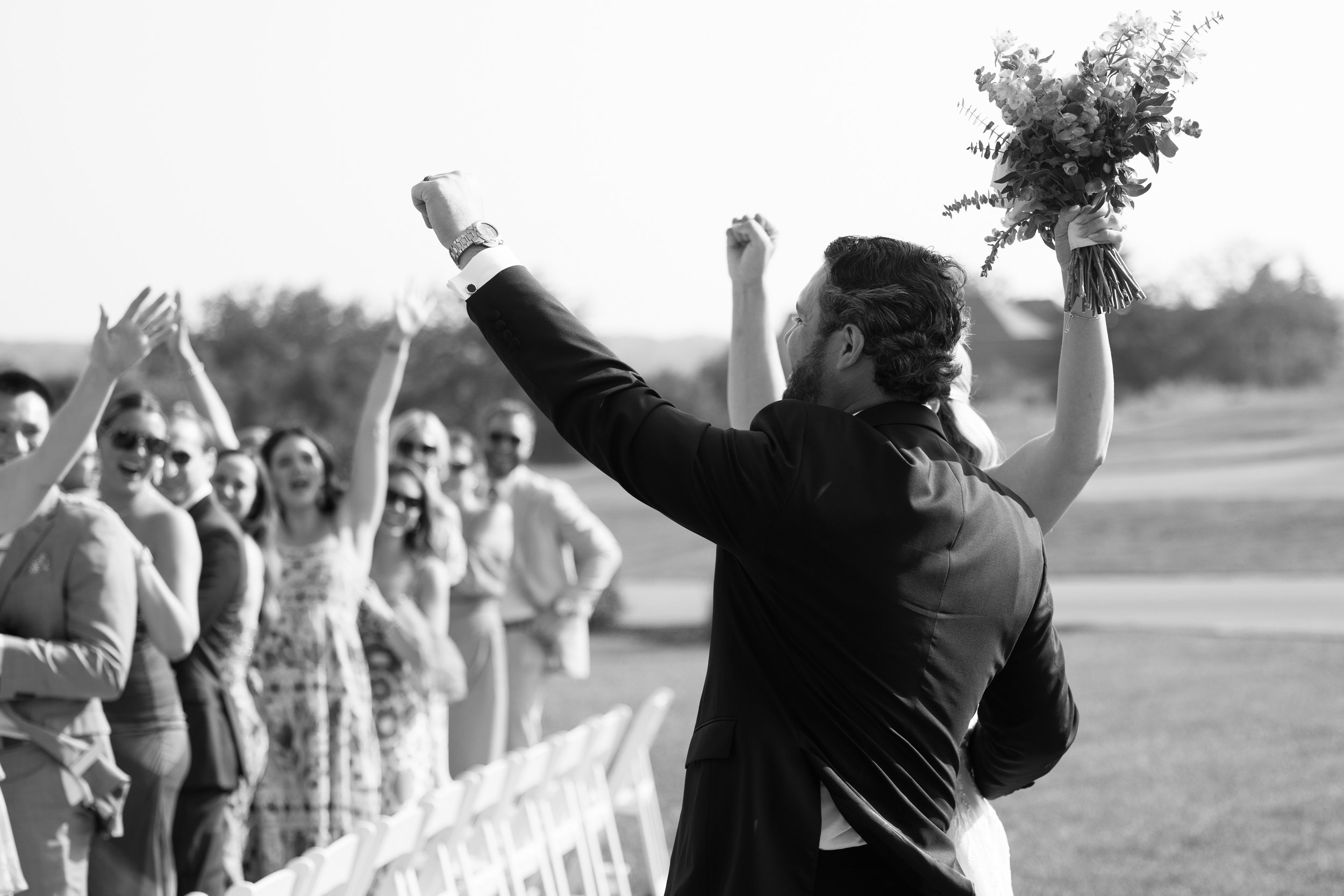 Man in a suit celebrating with a bouquet in hand at an outdoor wedding, surrounded by cheering guests.