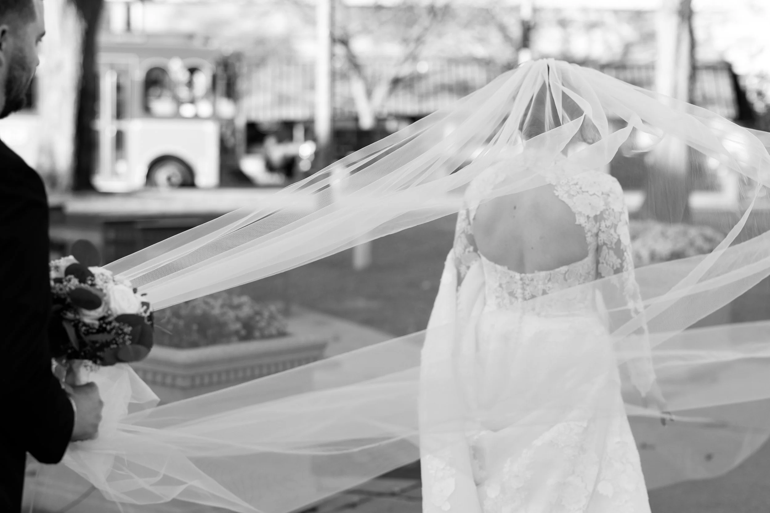 A bride in a lace wedding dress standing outdoors with a veil being held by a person on her left, partially covering her face and upper body.