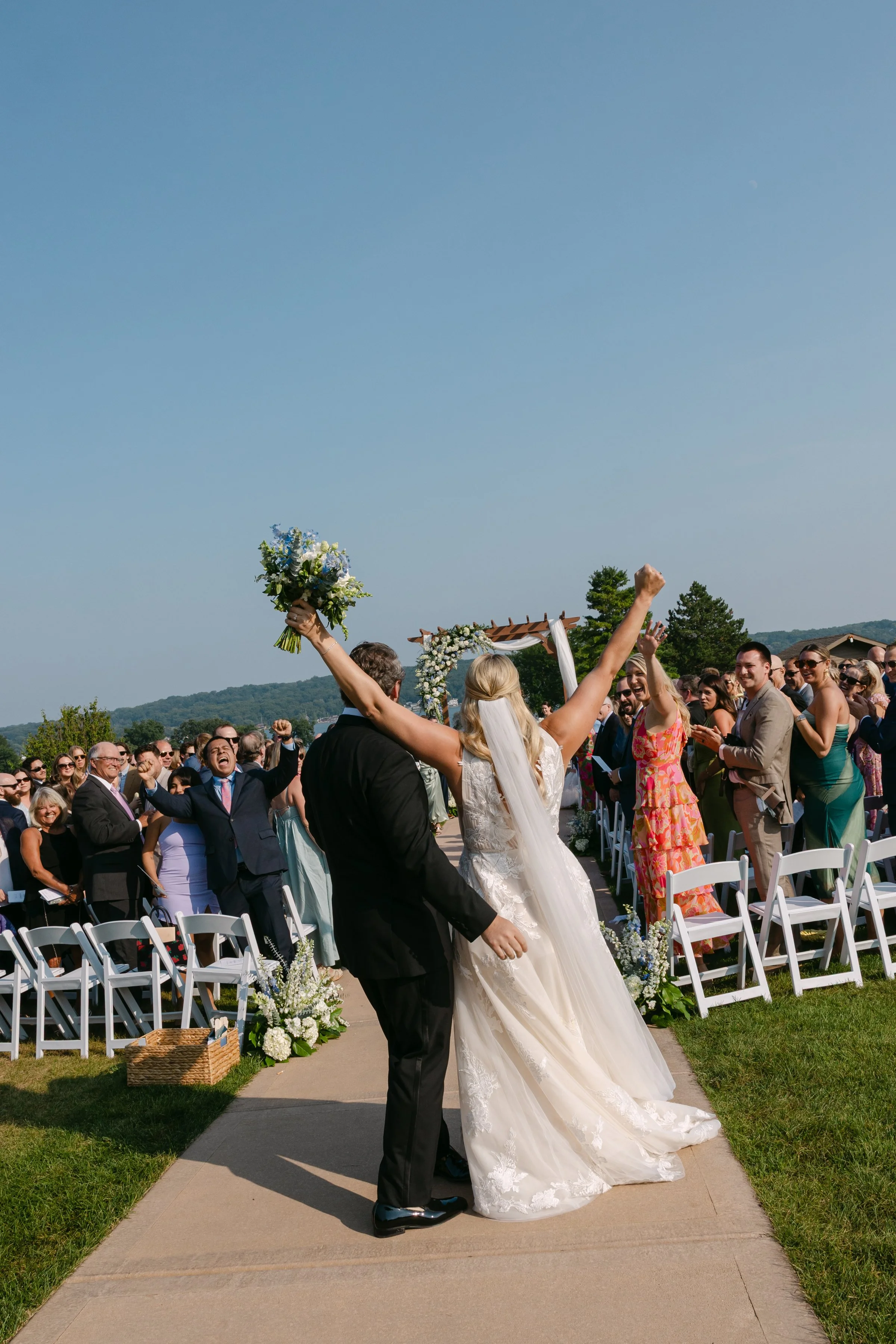 Wedding couple celebrating outdoors with guests cheering, bride holding a bouquet up, sunshine, chairs decorated with flowers, scenic background.