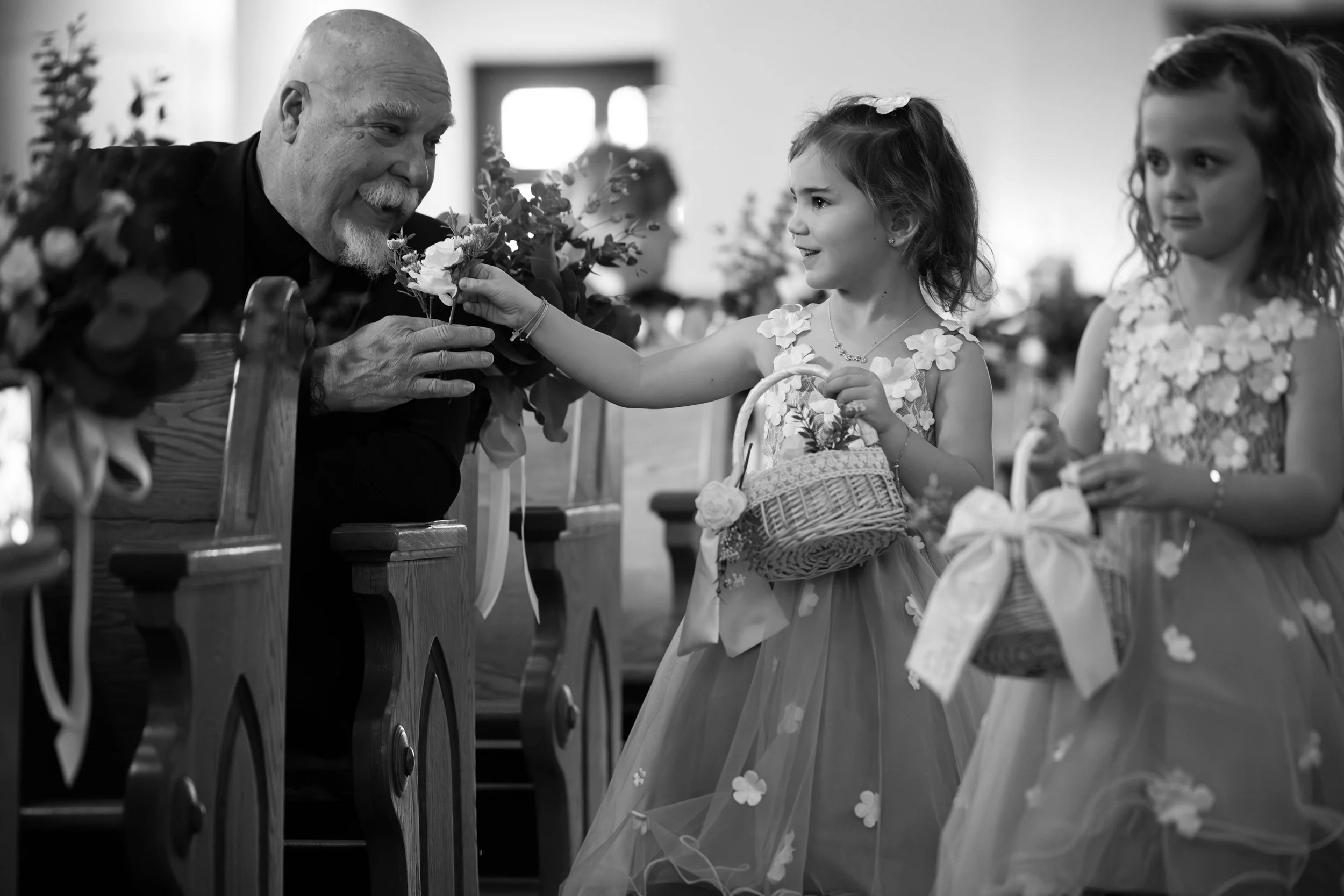 A young girl in a floral dress giving a flower to an older man sitting in a church pew, with another girl standing nearby holding a basket, both girls wearing similar dresses.