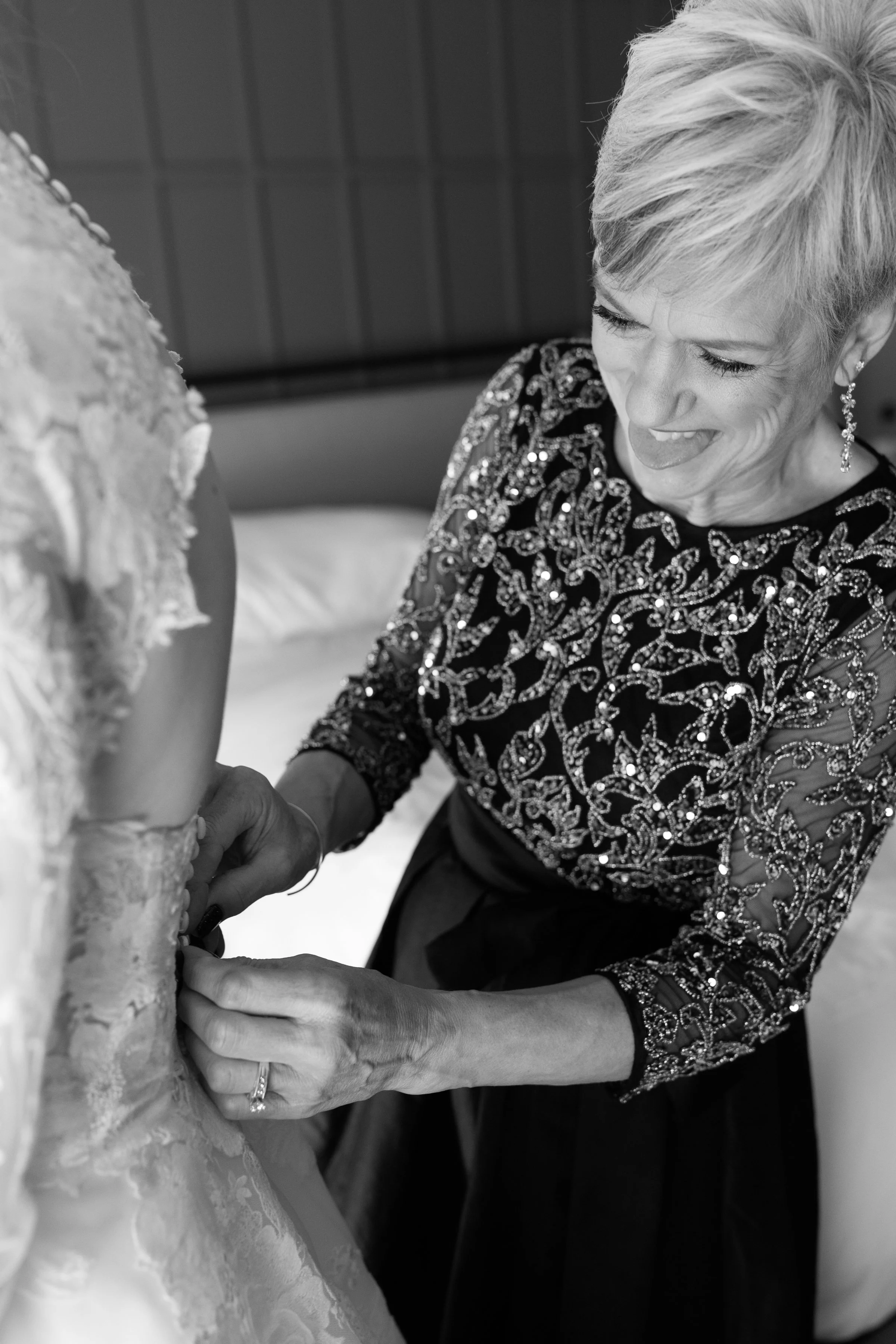 A woman, smiling and dressed in an ornate, sequin-embellished dress, helps fasten the back of a bride's wedding gown. The bride's gown features lace details, and the woman's cheerful expression indicates a joyful moment.