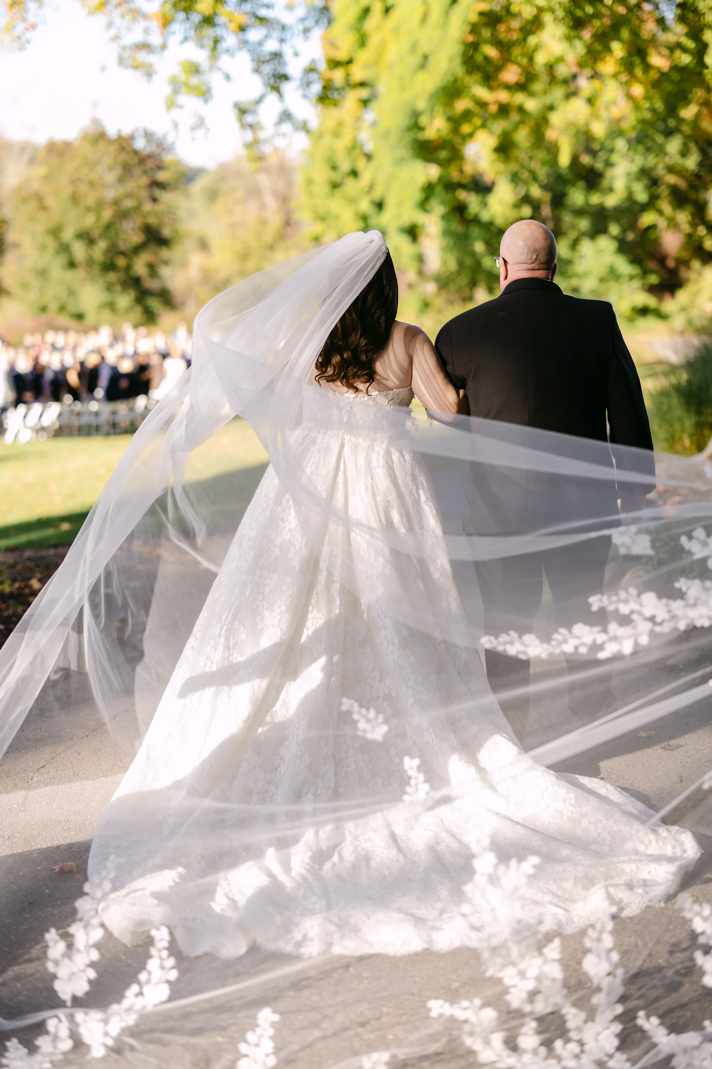 A bride and groom walking outdoors during their wedding ceremony, with guests seated in the background. The bride is wearing a white wedding gown with a long veil, and the groom is in a black suit. The trees around them are green and colorful, indica