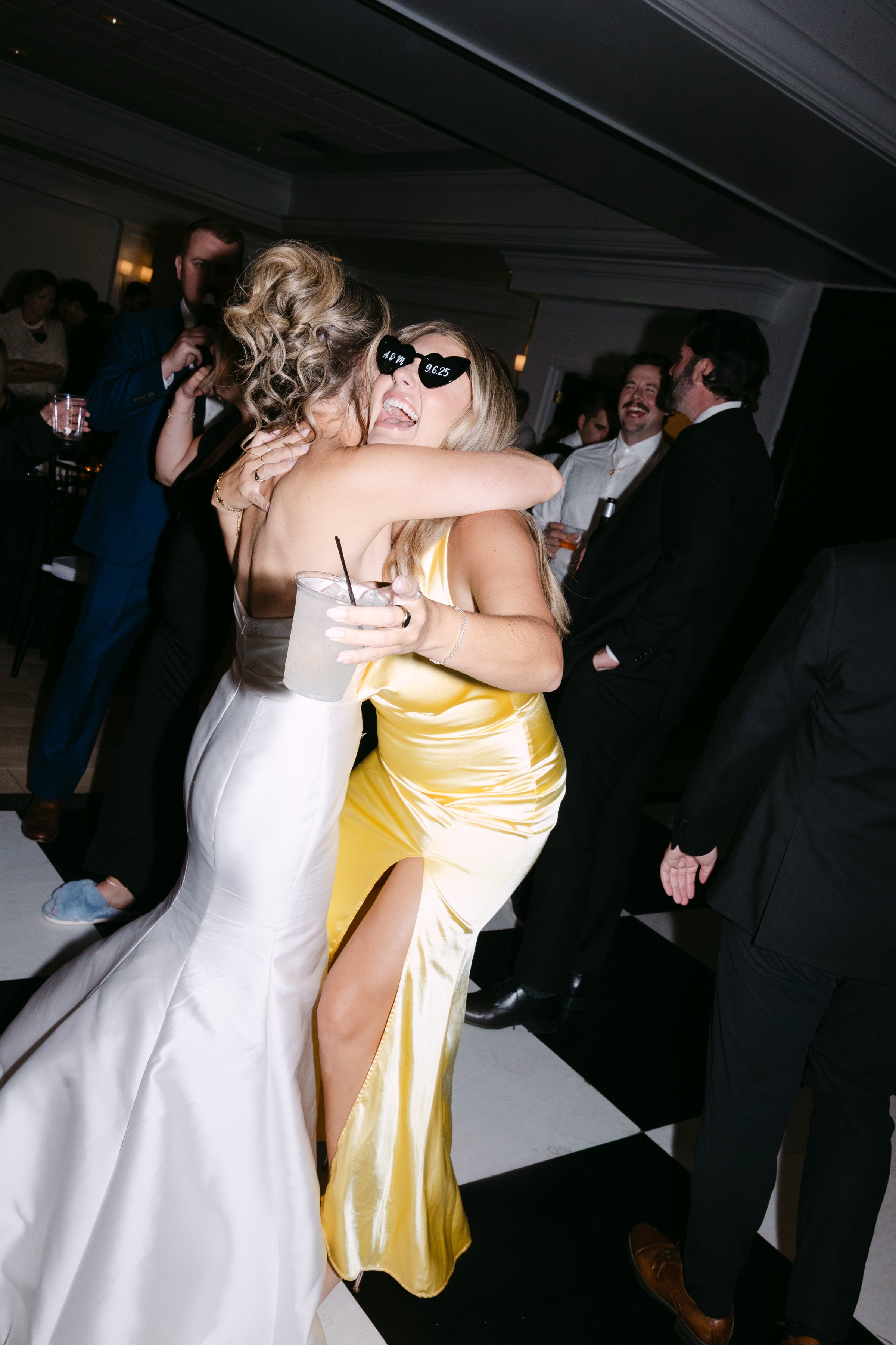 Two women embracing and celebrating at a party, one in a white dress and the other in a yellow satin dress, holding a drink, with other people dancing and socializing in the background.