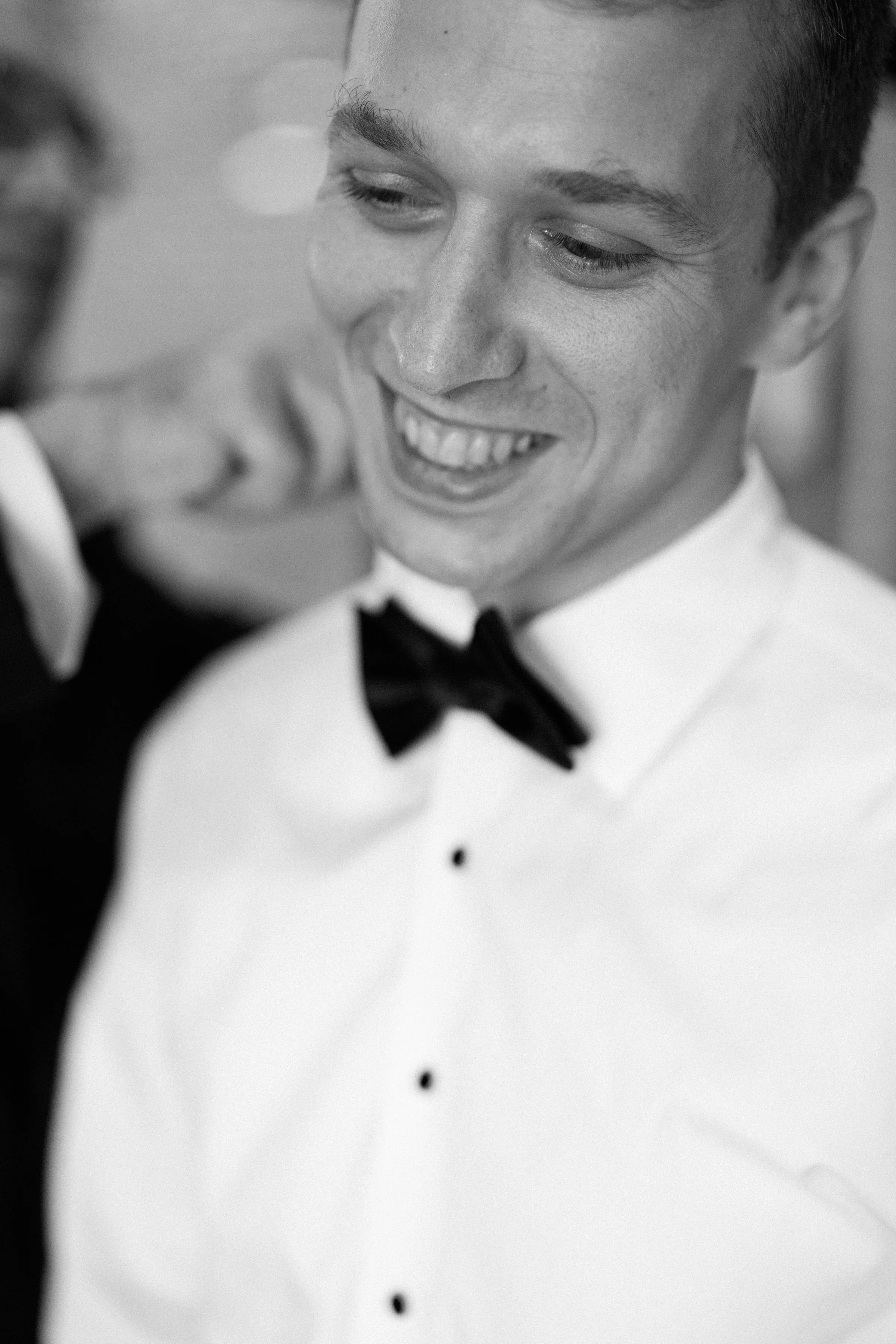 A smiling young man in a tuxedo with a black bow tie, having his collar adjusted by another person, in black and white photo.
