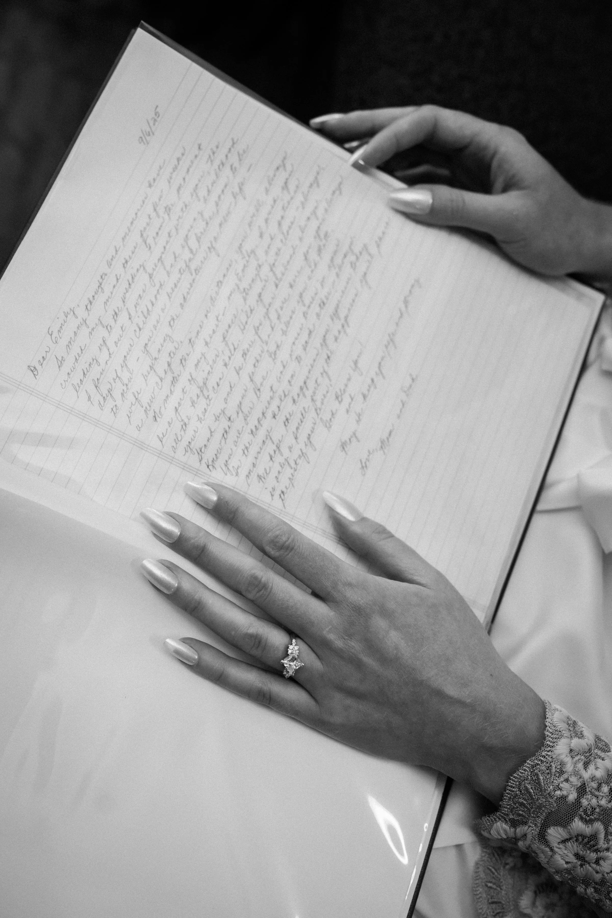 A woman with manicured nails and a wedding ring reads a handwritten letter in a notebook on a table.