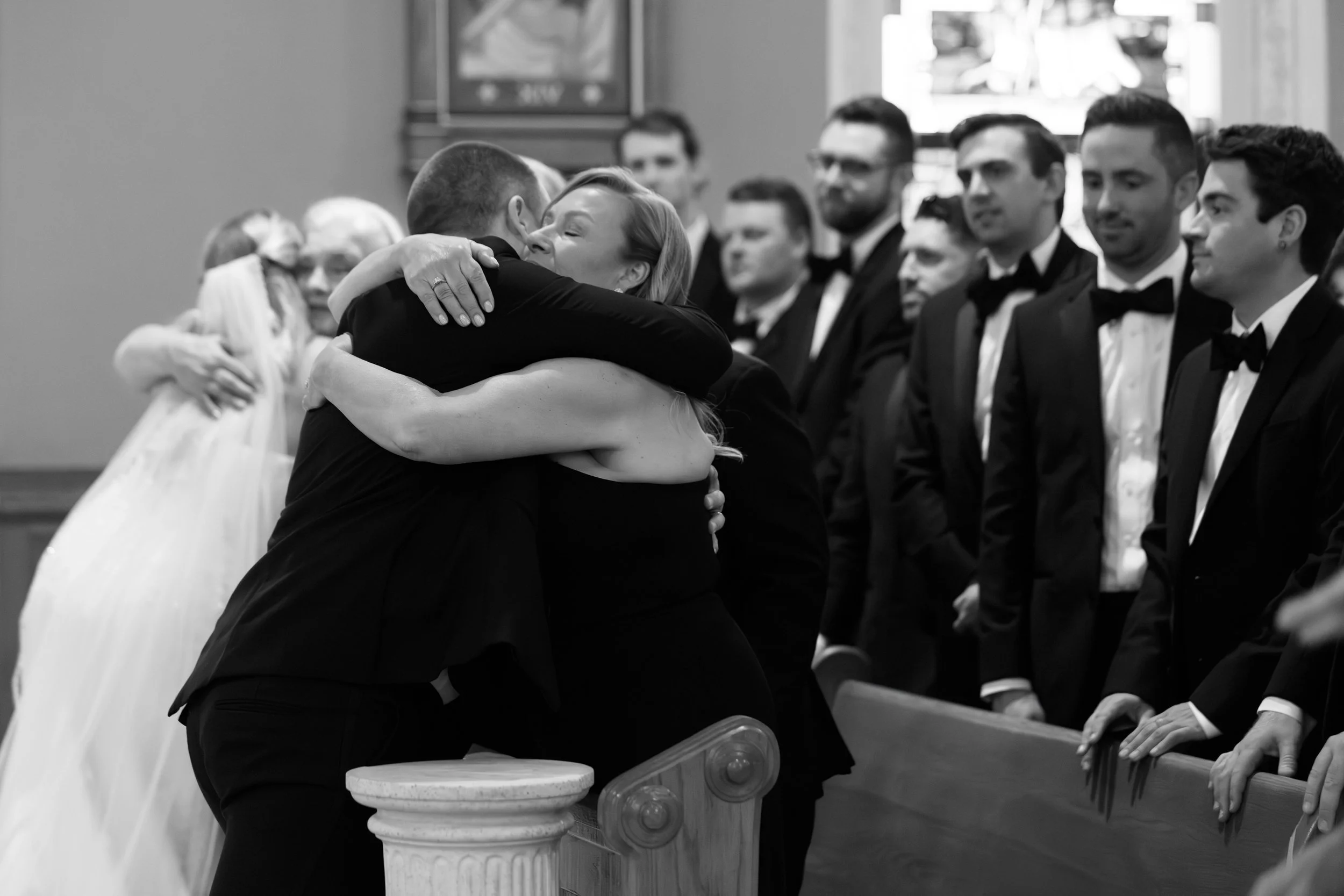 Two women hugging and embracing at a formal gathering, with a line of men in tuxedos watching in a church or ceremony setting.