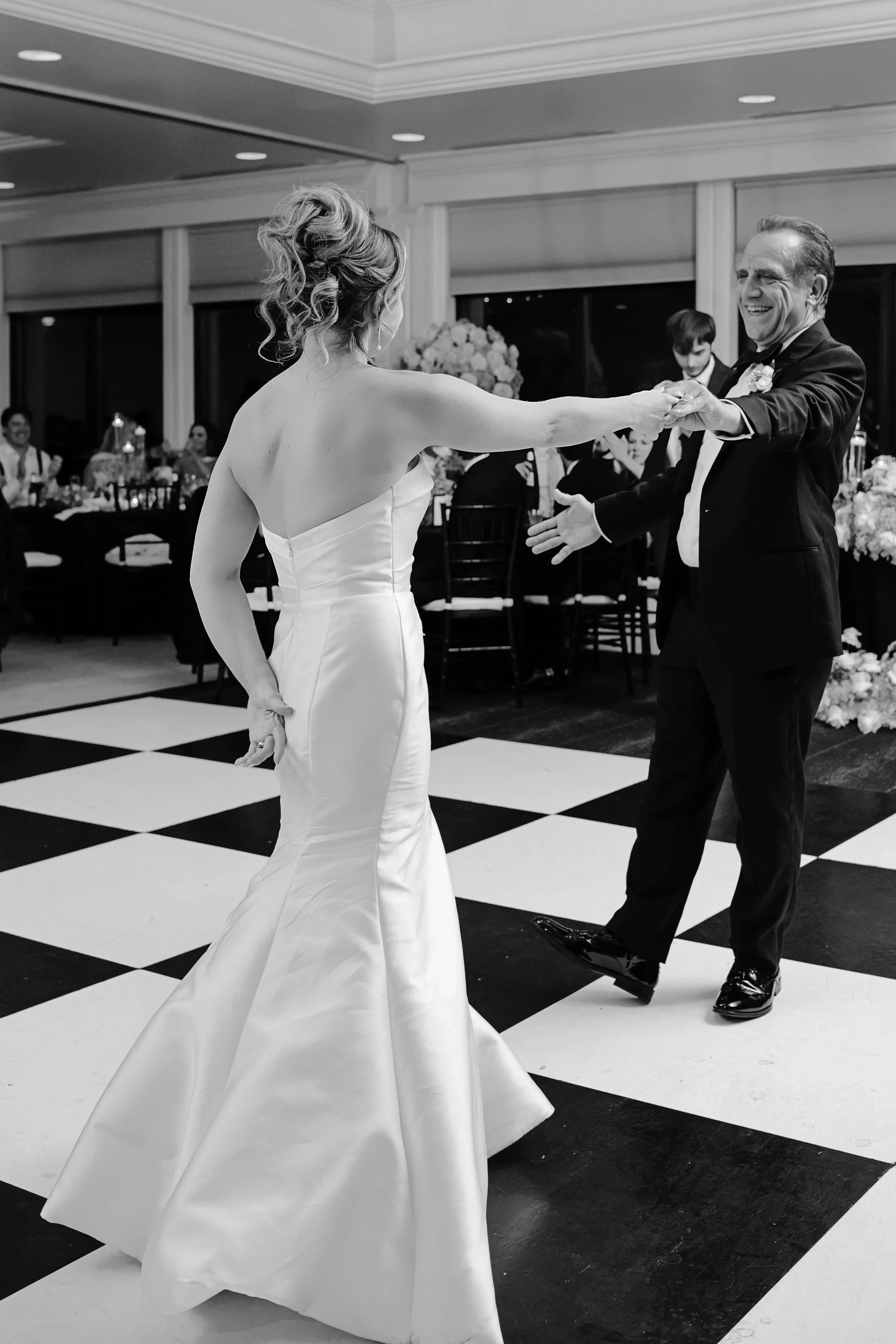 A bride and groom dancing at their wedding reception, with guests seated at tables in the background.