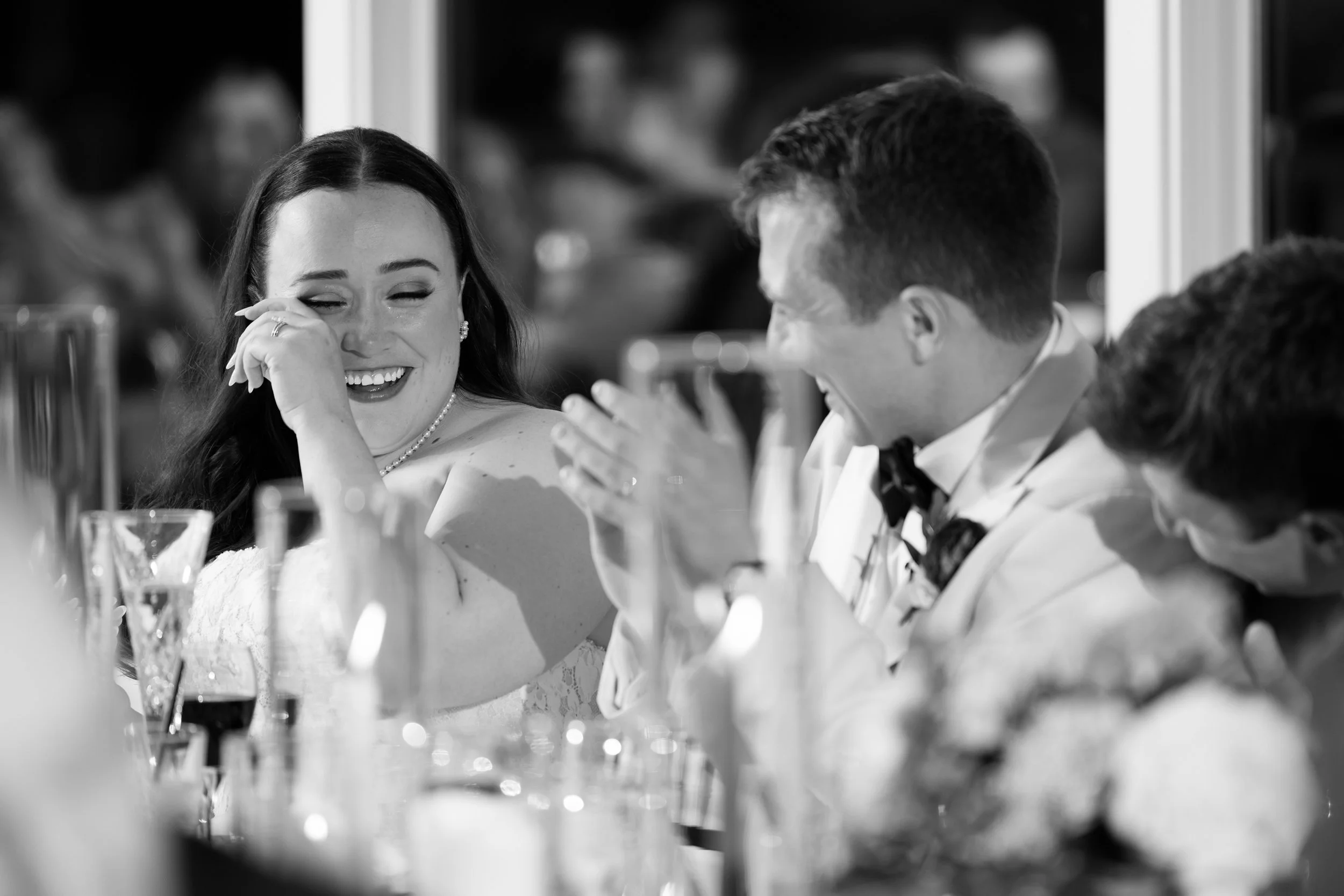 Black and white photo of a woman laughing and wiping a tear from her eye while sitting at a formal event with men in tuxedos.