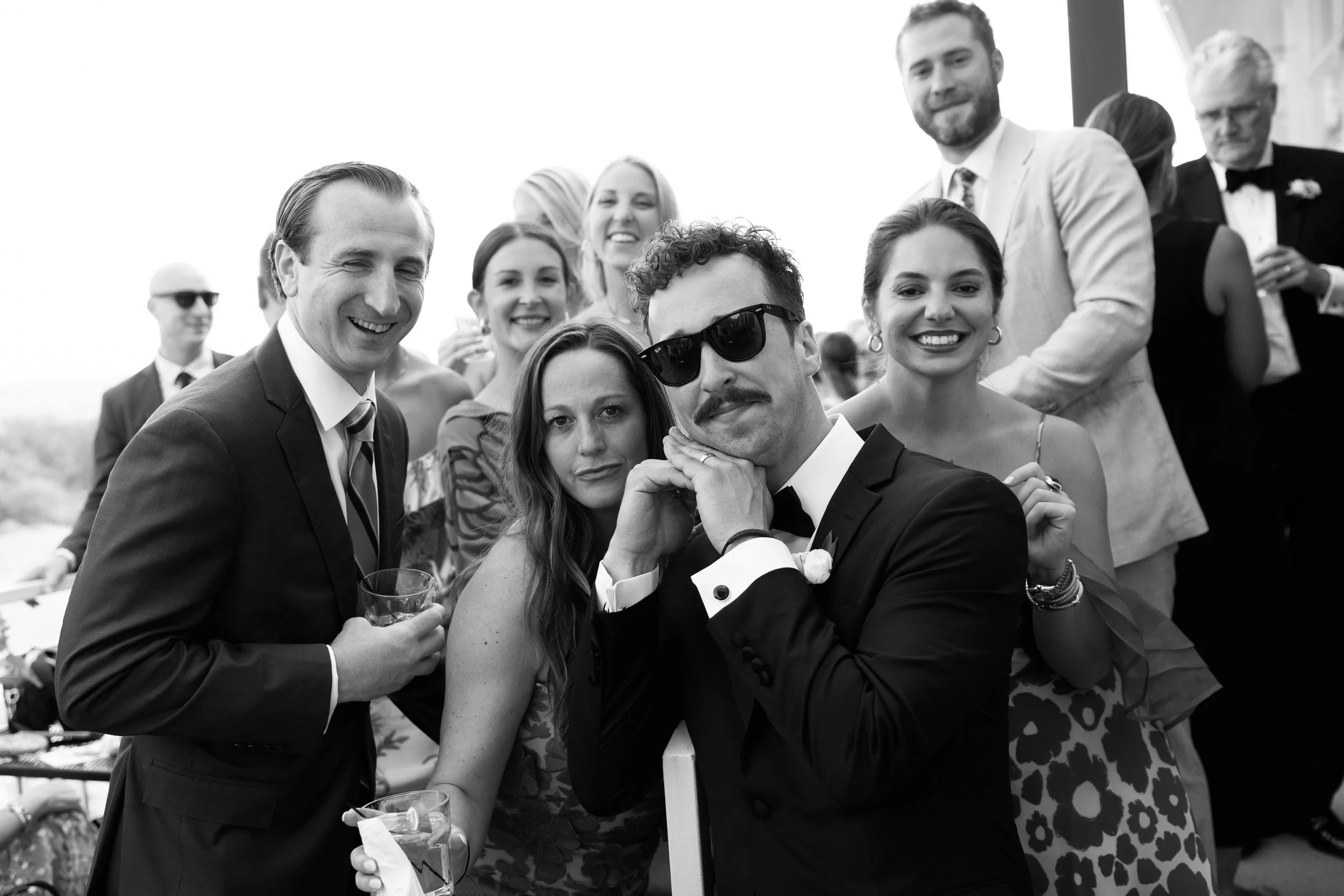Group of people at a social gathering or celebration, mostly smiling and looking at the camera, dressed in formal attire, outdoors with a bright sky.
