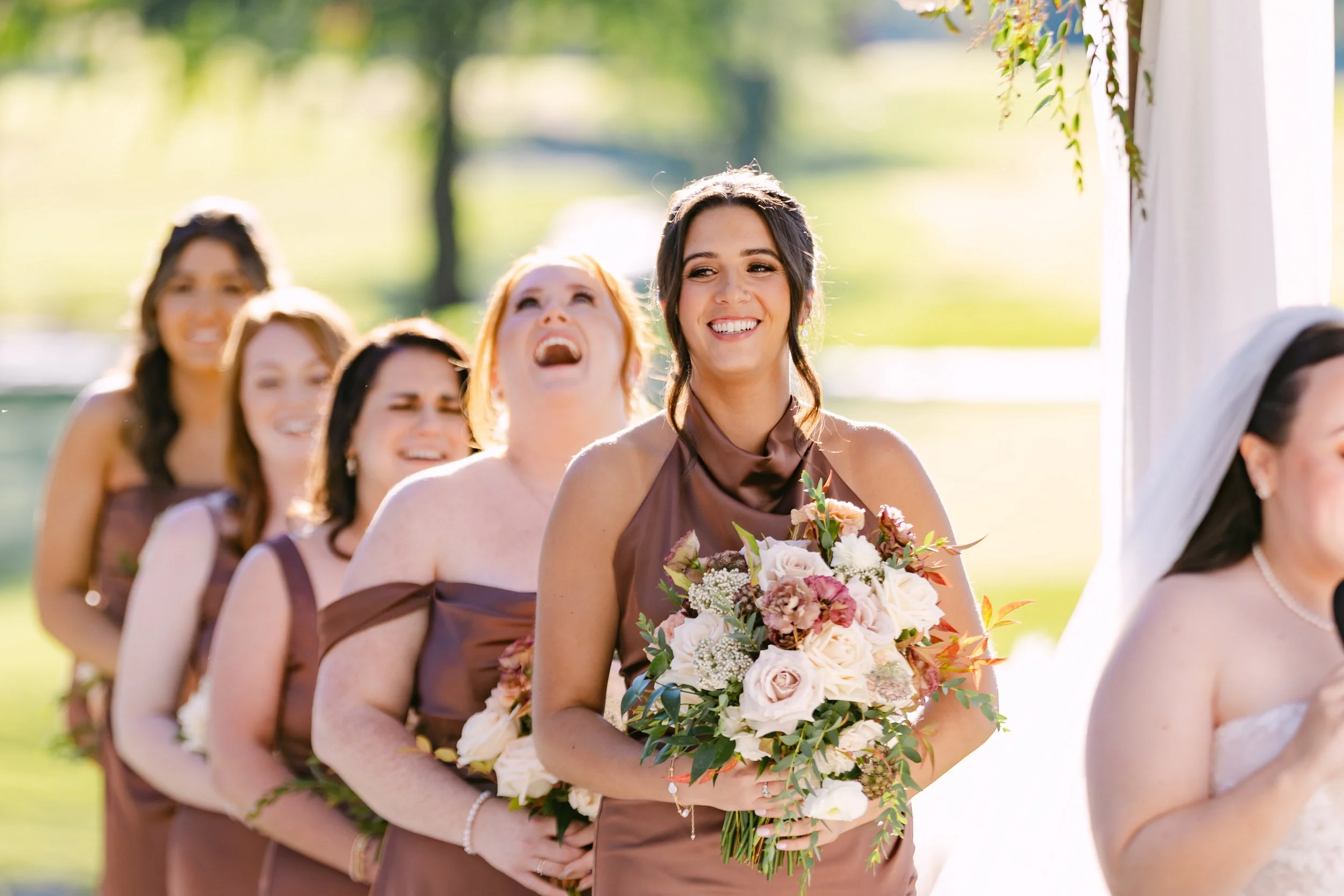 A smiling bride holding a bouquet of flowers at an outdoor wedding ceremony, standing with her bridesmaids also holding bouquets and dressed in similar brown dresses, with greenery and sunlight in the background.