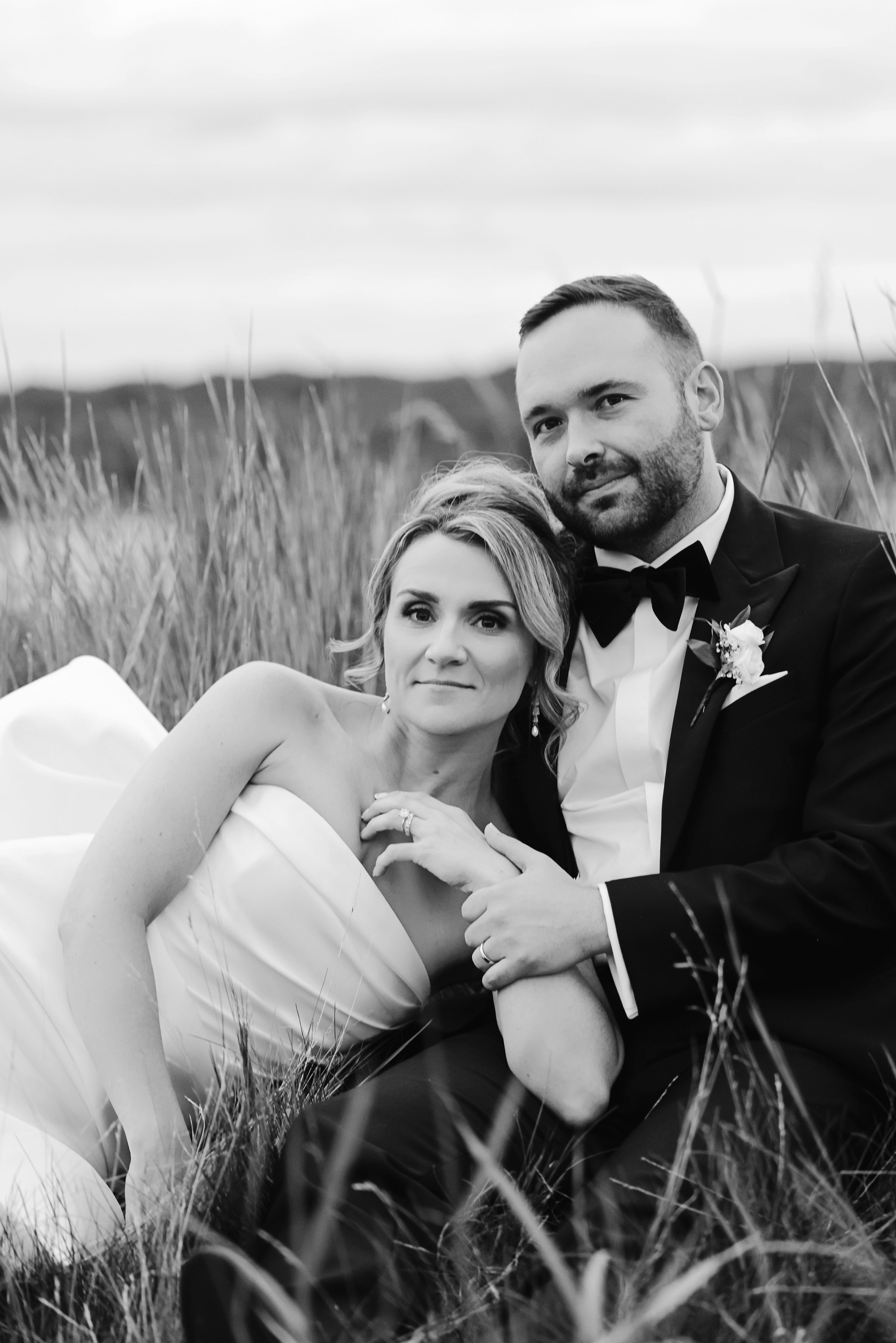 Black and white photograph of a newlywed couple sitting in a grassy field. The woman is reclining on the grass, wearing a strapless wedding dress, and the man is sitting behind her, dressed in a tuxedo with a bow tie, gently holding her hand and looking at the camera.
