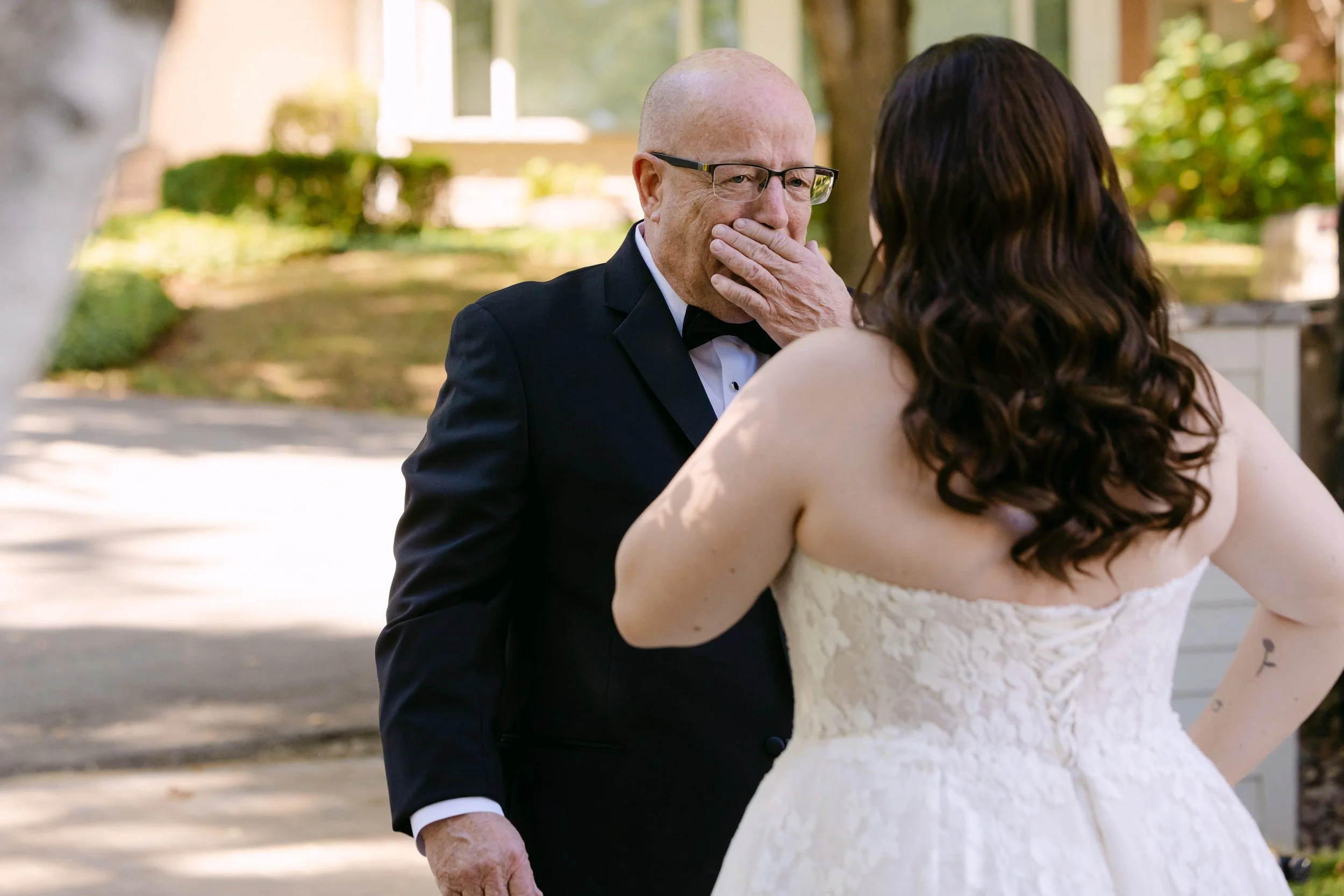 A bride and an older man in a tuxedo are in an outdoor setting. The man has his hand over his mouth, and the bride is facing him with her back to the camera, touching his face.