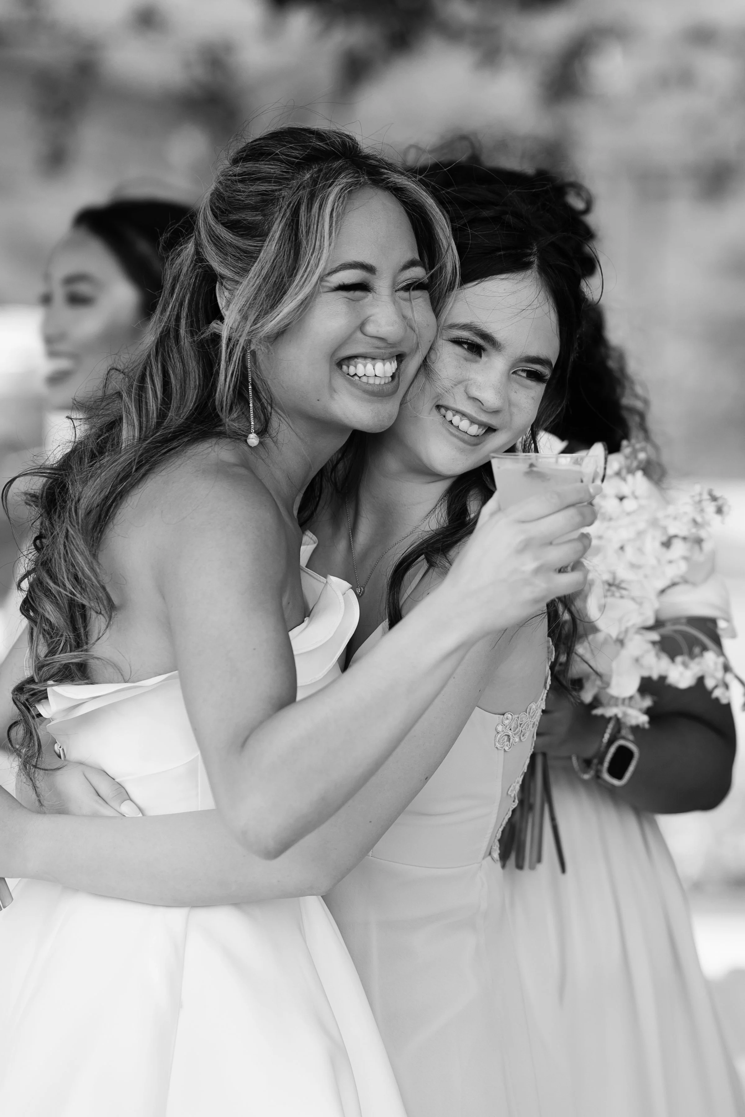 Two women in wedding dresses smiling and hugging, one holding a cocktail, at a wedding celebration.