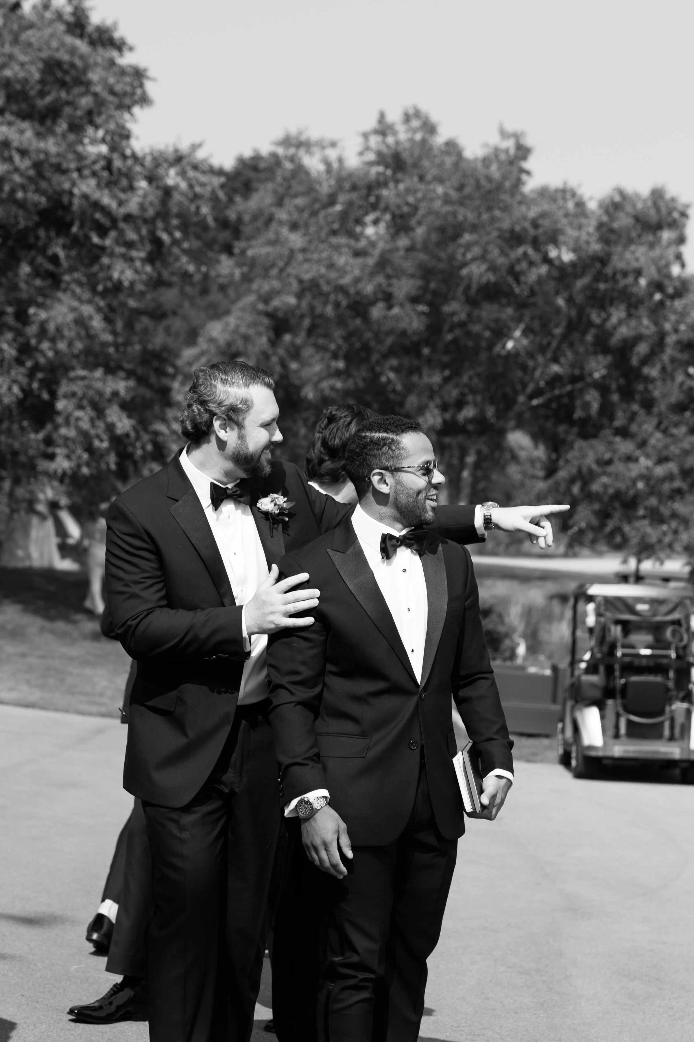 Three men dressed in tuxedos, one of them pointing, standing outdoors with trees in the background.