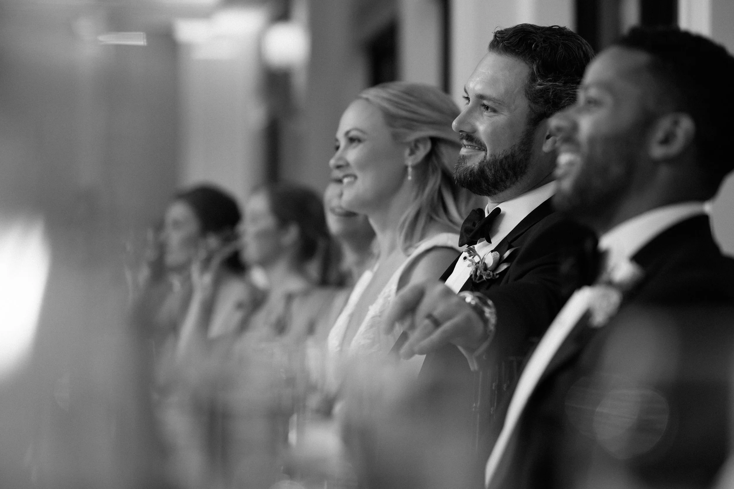 Black and white photo of wedding reception with guests in formal attire, sitting and smiling in a row.
