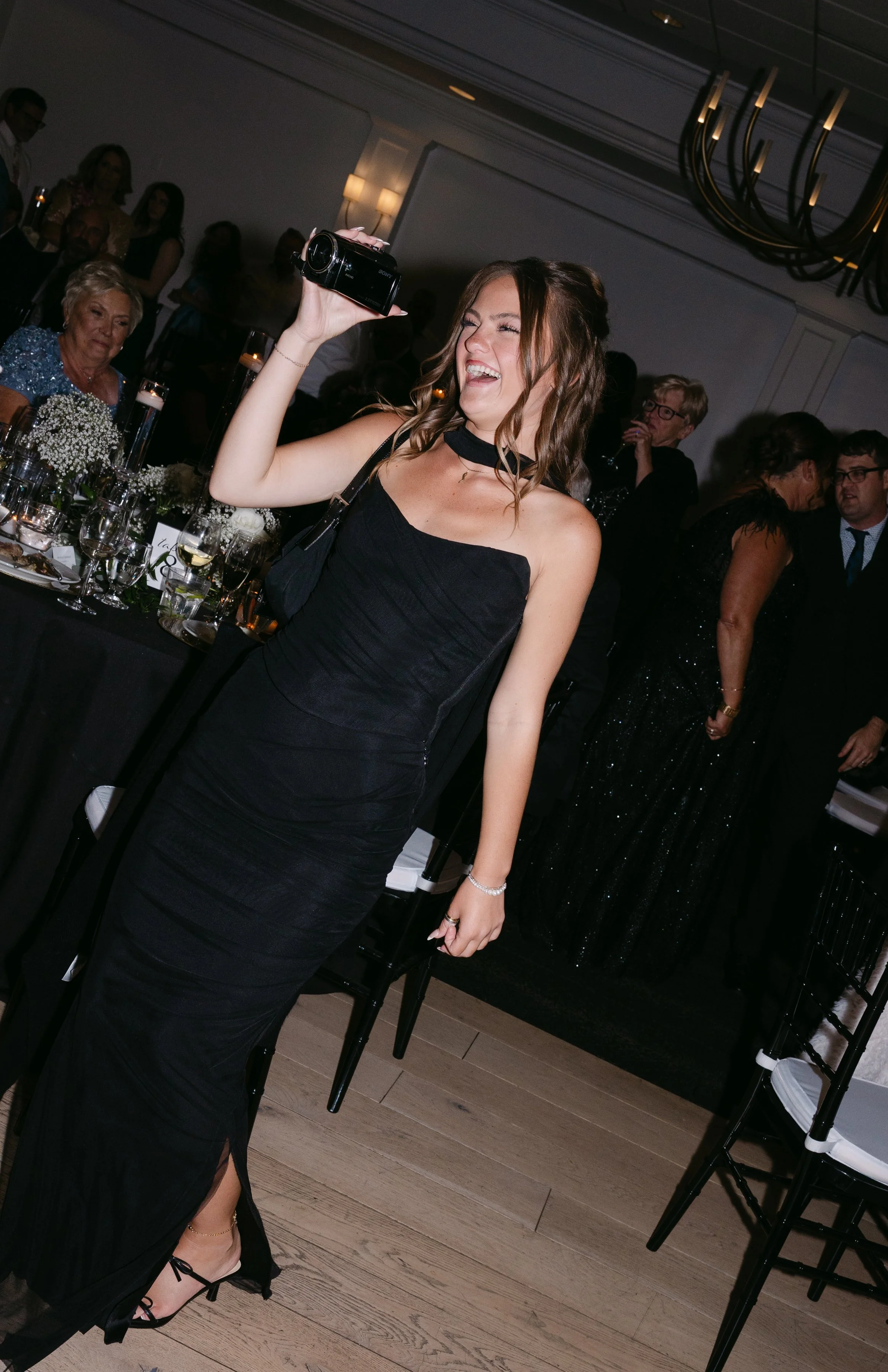 A young woman wearing a black evening gown, smiling and taking a photo with a camera at a formal event or wedding reception.