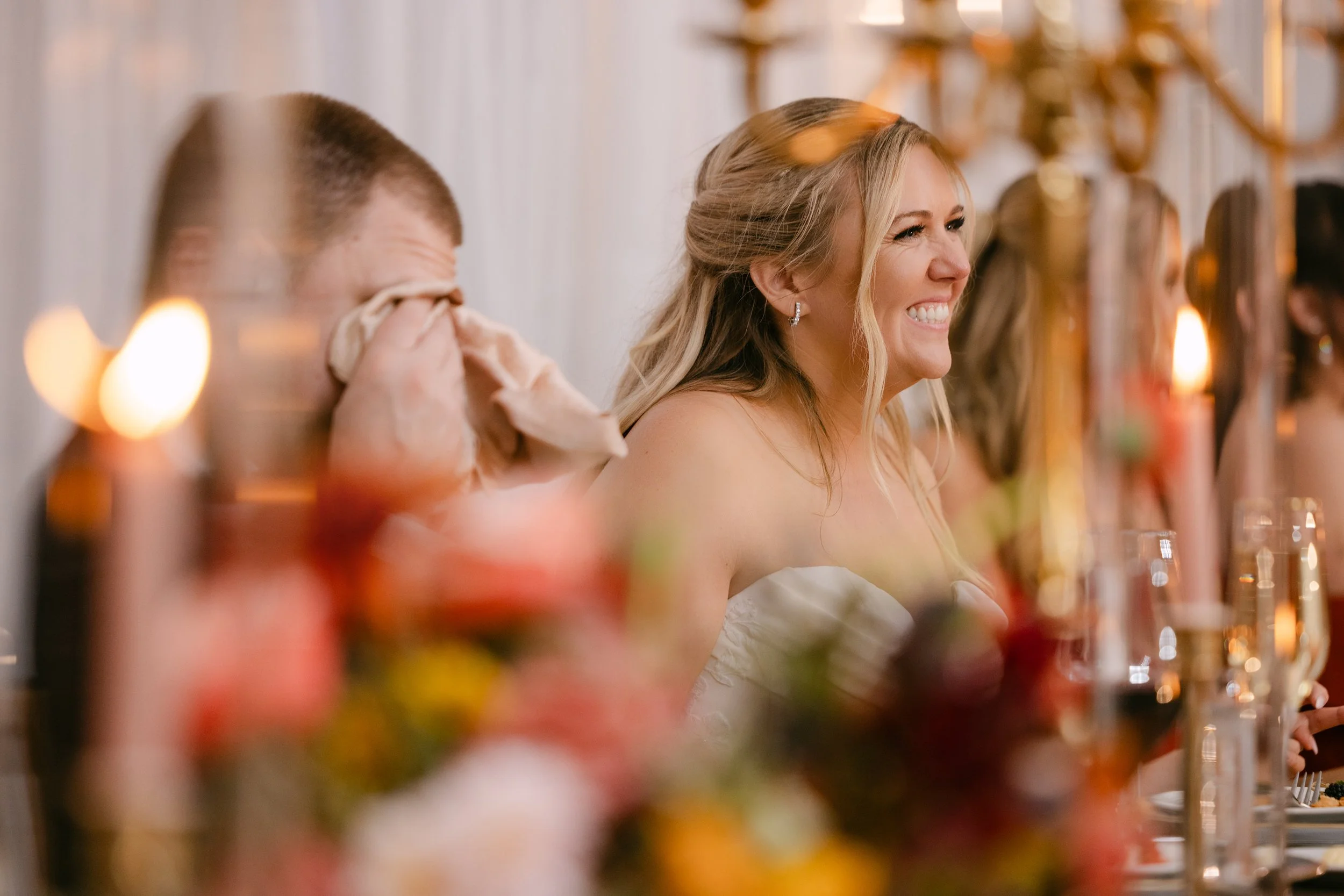 A woman is smiling and appears emotional, sitting at a formal event or wedding. She has long, wavy blonde hair and is wearing earrings and a strapless dress. Other people are visible in the background, and the table is decorated with flowers and cand