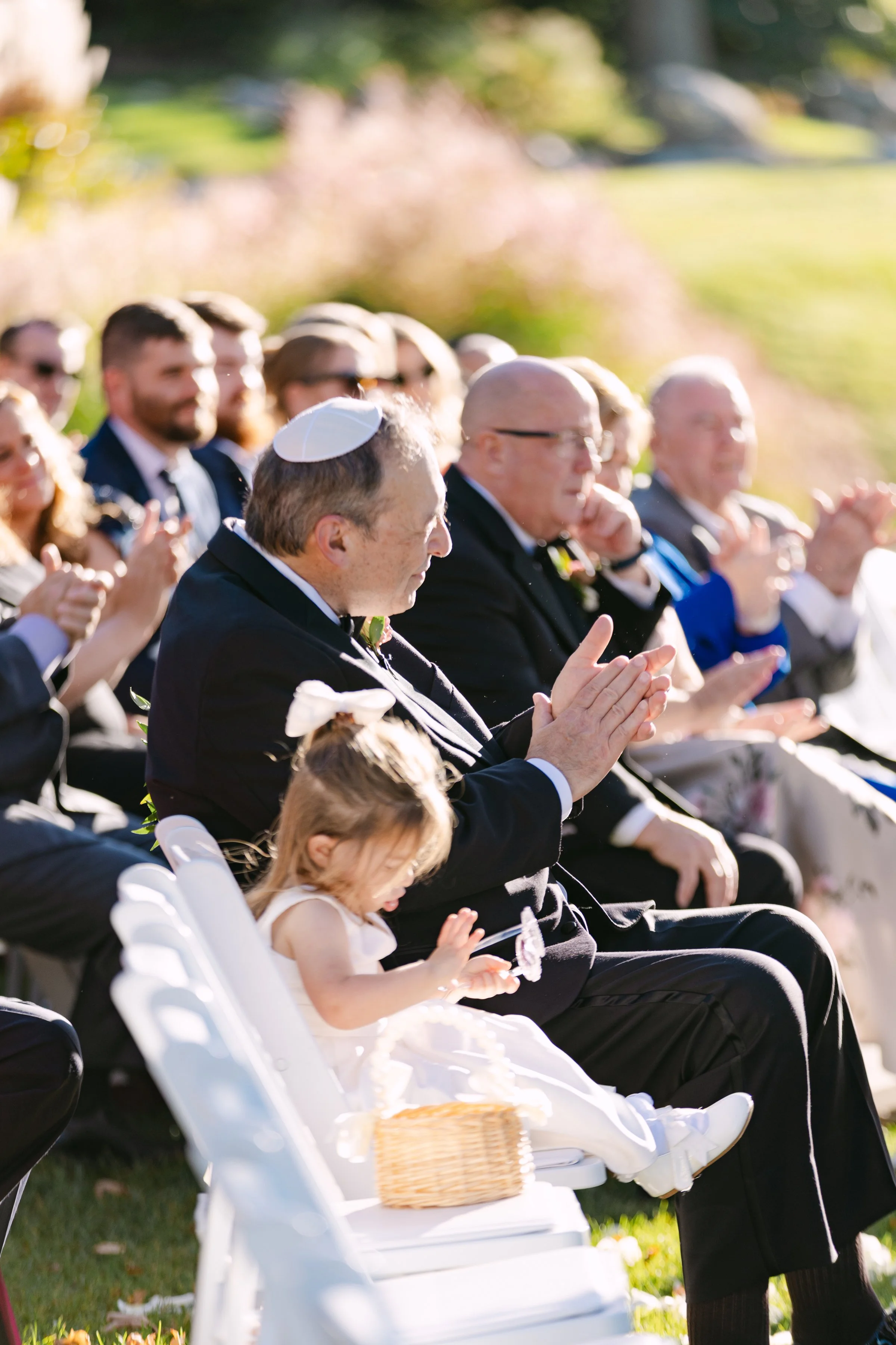 People seated outdoors at a wedding ceremony, clapping, with a young girl in a white dress sitting on a white chair, holding a basket, and a man wearing a kippah.