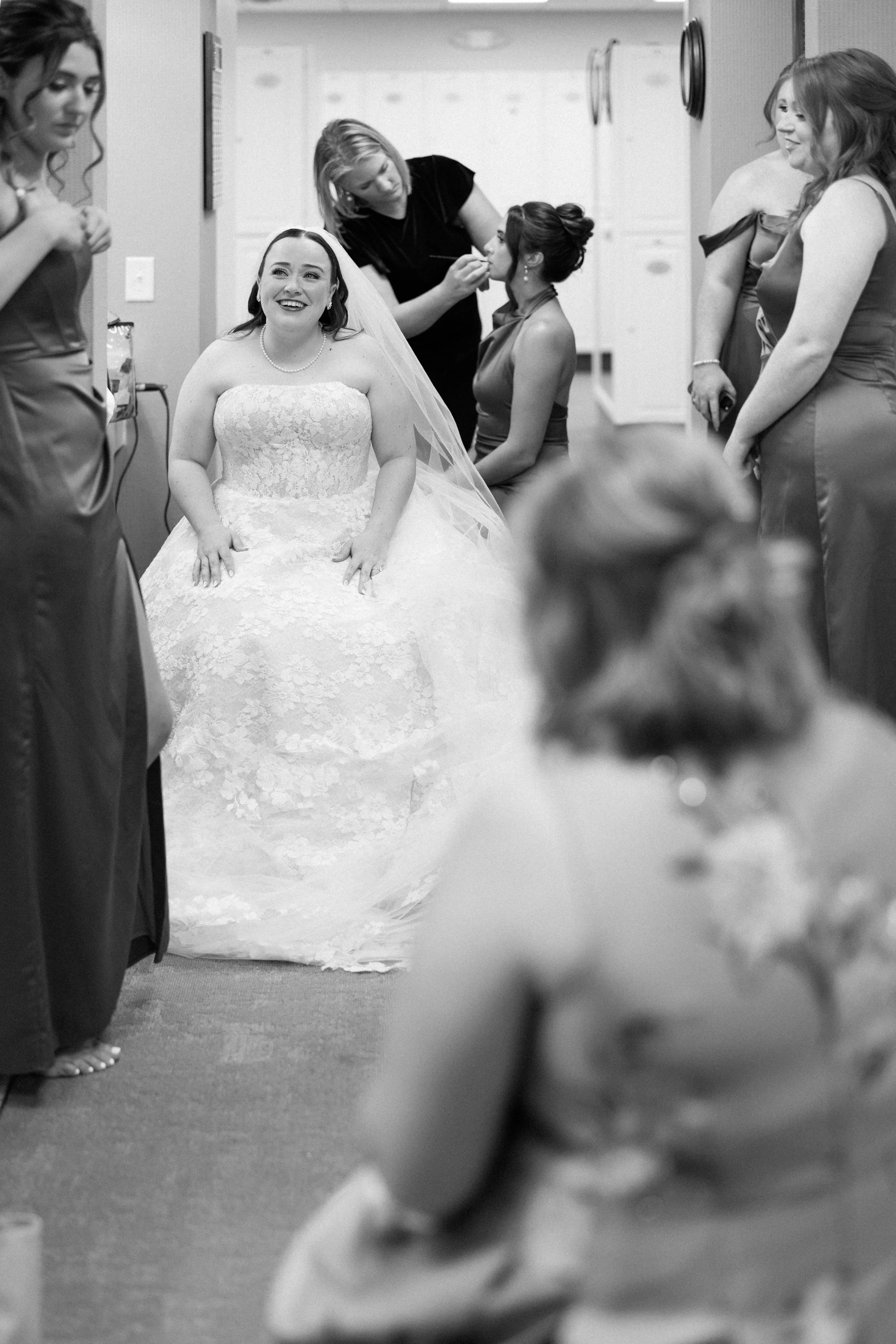 A bride sitting in a room surrounded by bridesmaids getting her makeup and hair done, smiling