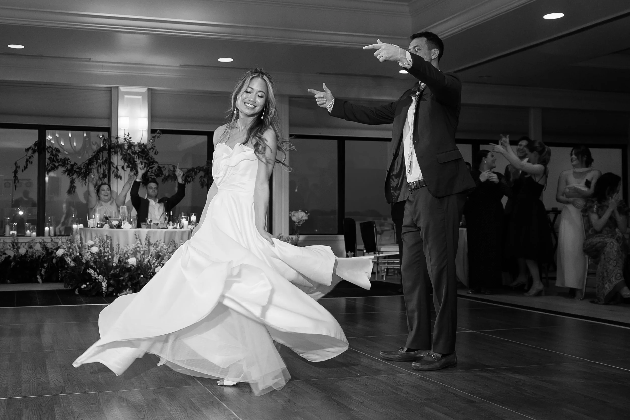 A bride in a white wedding dress dancing in a wedding reception hall while a man in a suit points and dances with her. Several guests are in the background, some clapping and smiling.