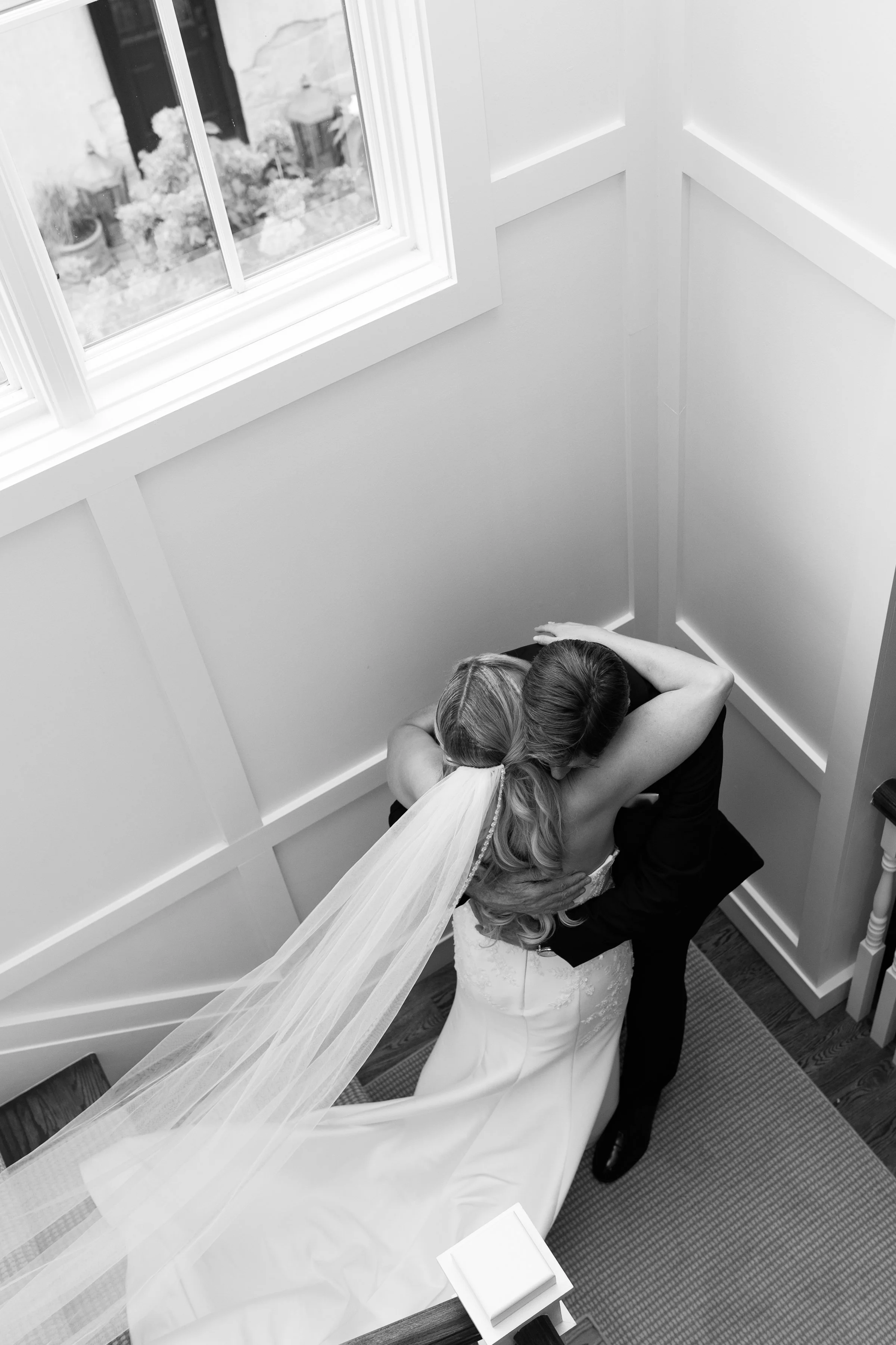 A bride in a wedding gown and veil hugging a man, likely her groom, in a stairwell near a window.