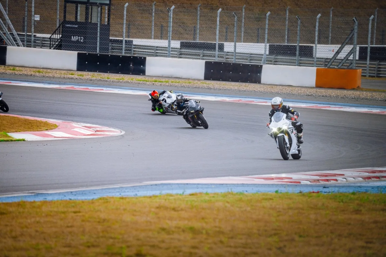 Motorcycle racers leaning into a curve on a racetrack, with the lead rider on a white motorcycle and others following behind.