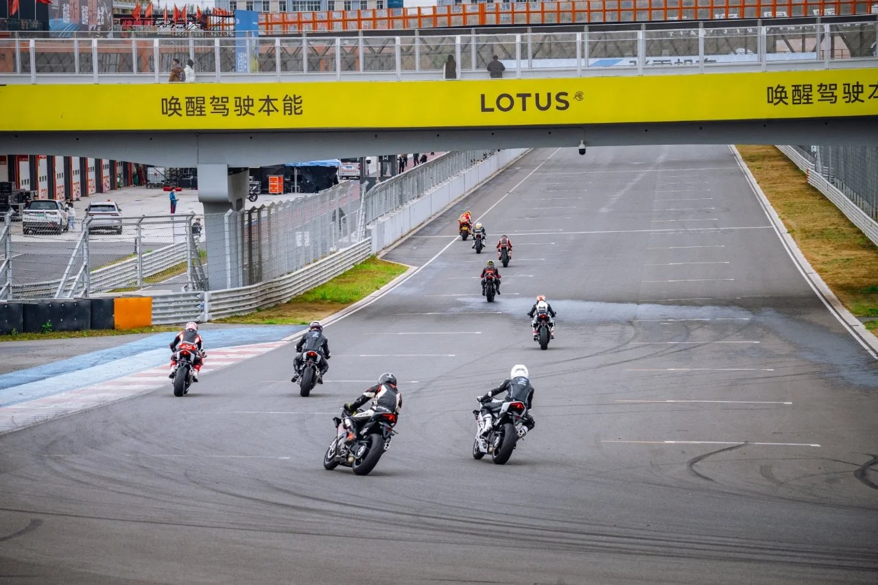Multiple motorcyclists racing on a track, leaning into a turn at a race circuit with grandstands and a bridge with a yellow banner.