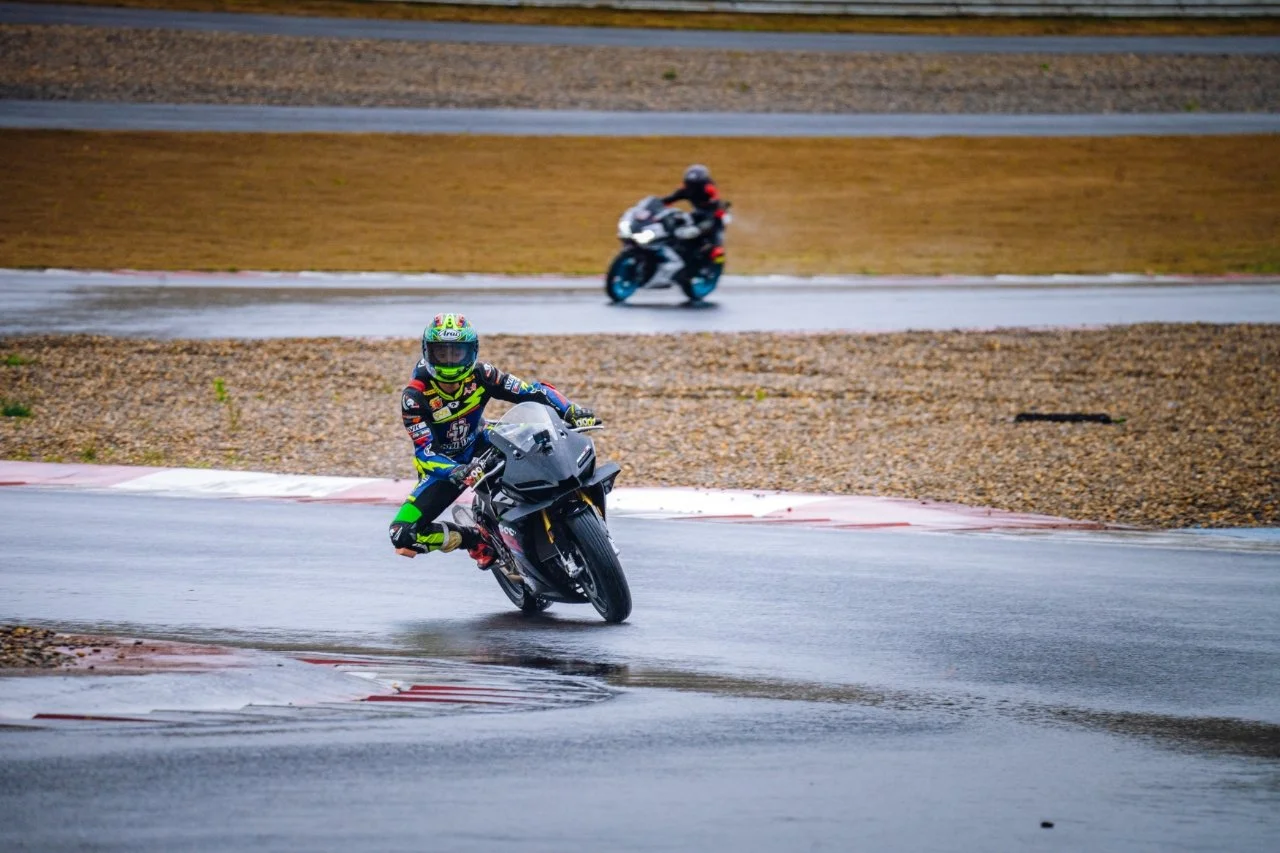 Two motorcyclists riding on a wet race track during a rainy day, the one in the front leaning into a turn and the other farther back on the track.