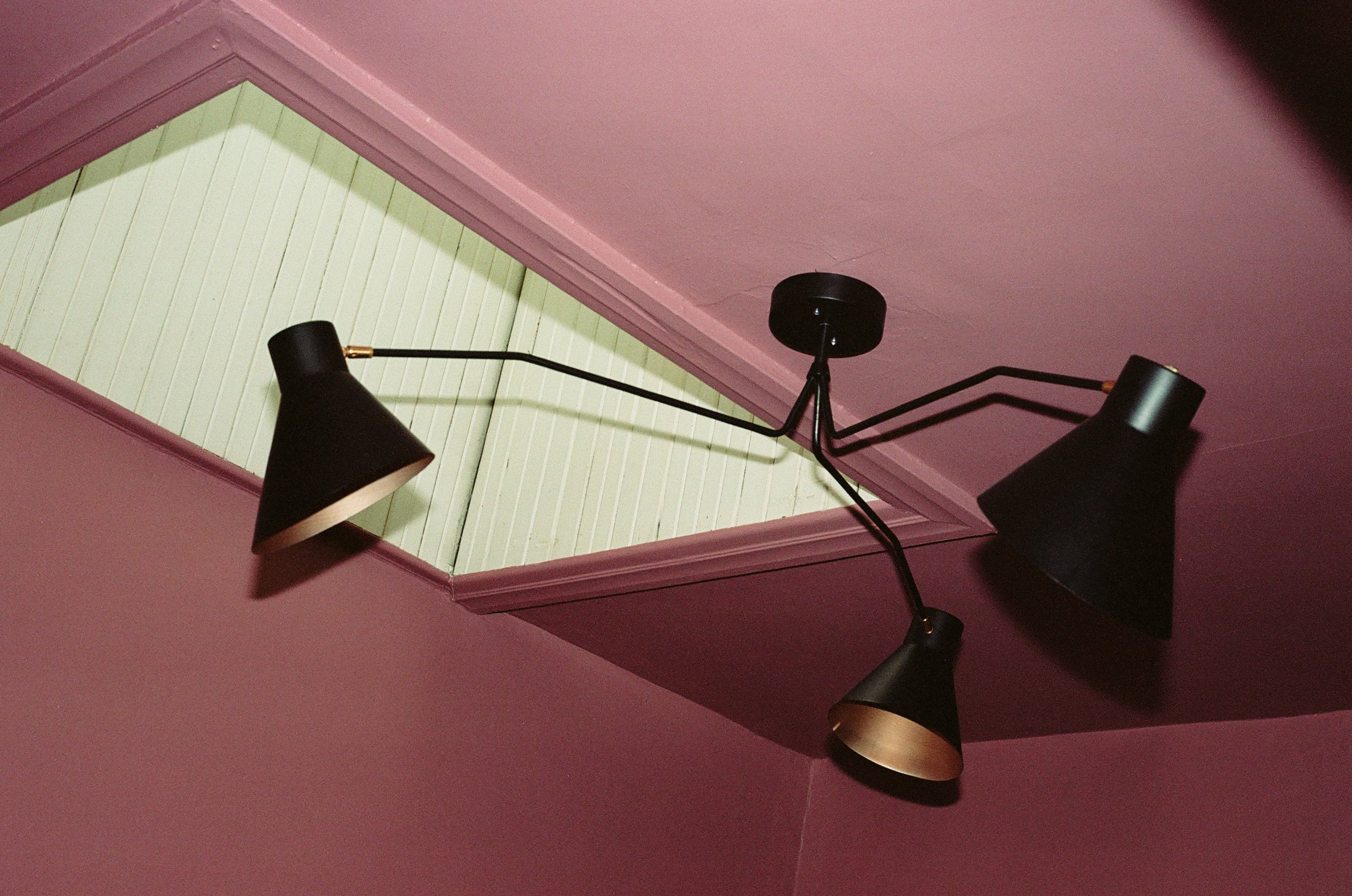 Détail luminaire suspendu noir à trois branches sous une lucarne, plafond rose