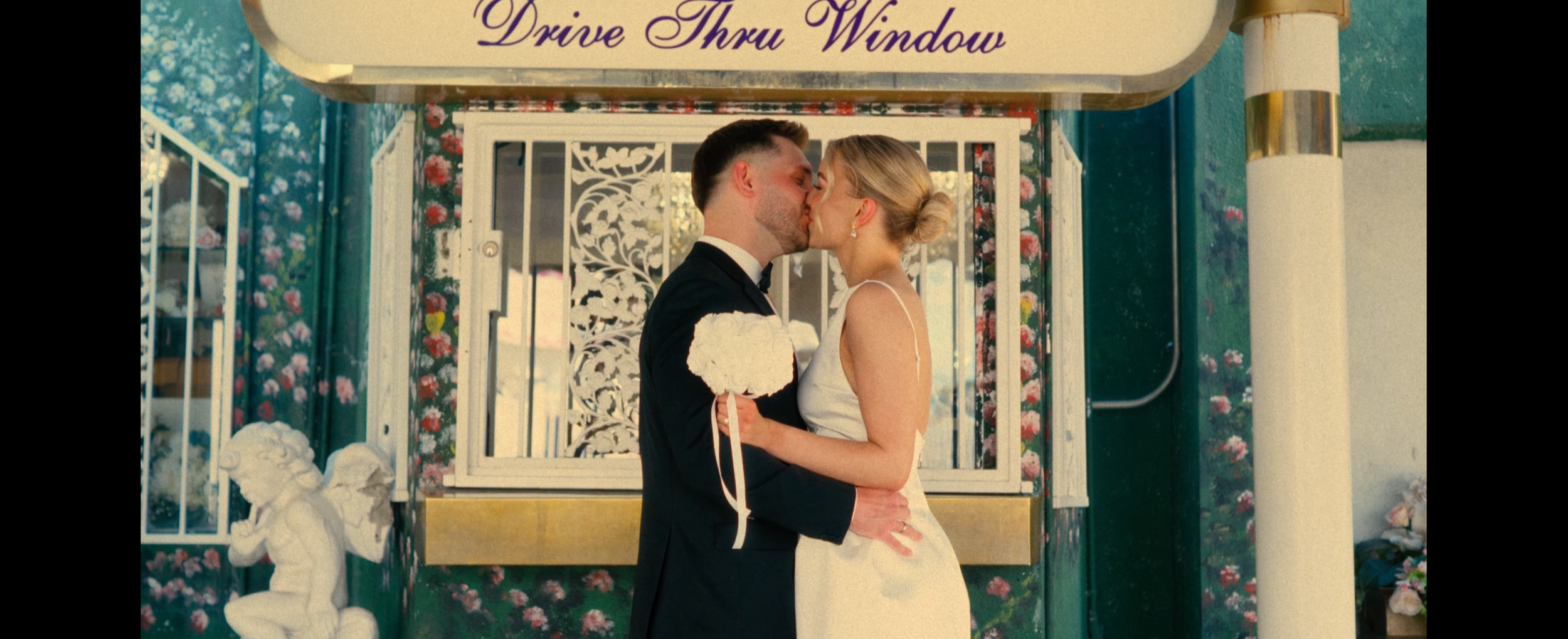 A couple in wedding attire sharing a kiss in front of a drive-thru window with floral decor.