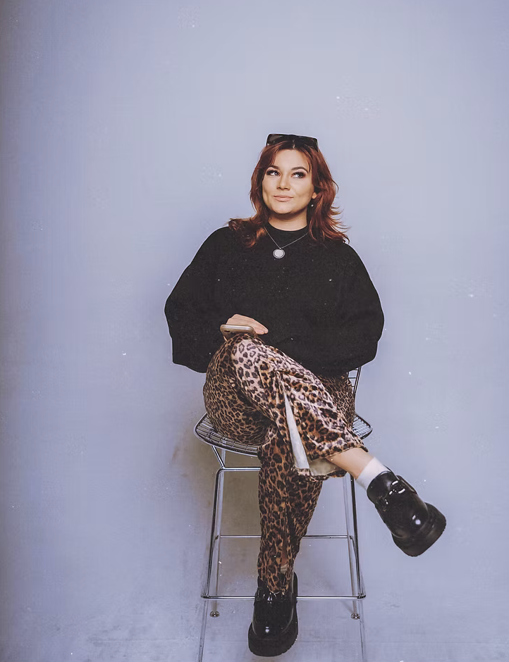 A woman with red hair and sunglasses on her head, sitting on a metal chair against a plain background, wearing a black sweater, leopard print pants, and black platform shoes, looking to the side.